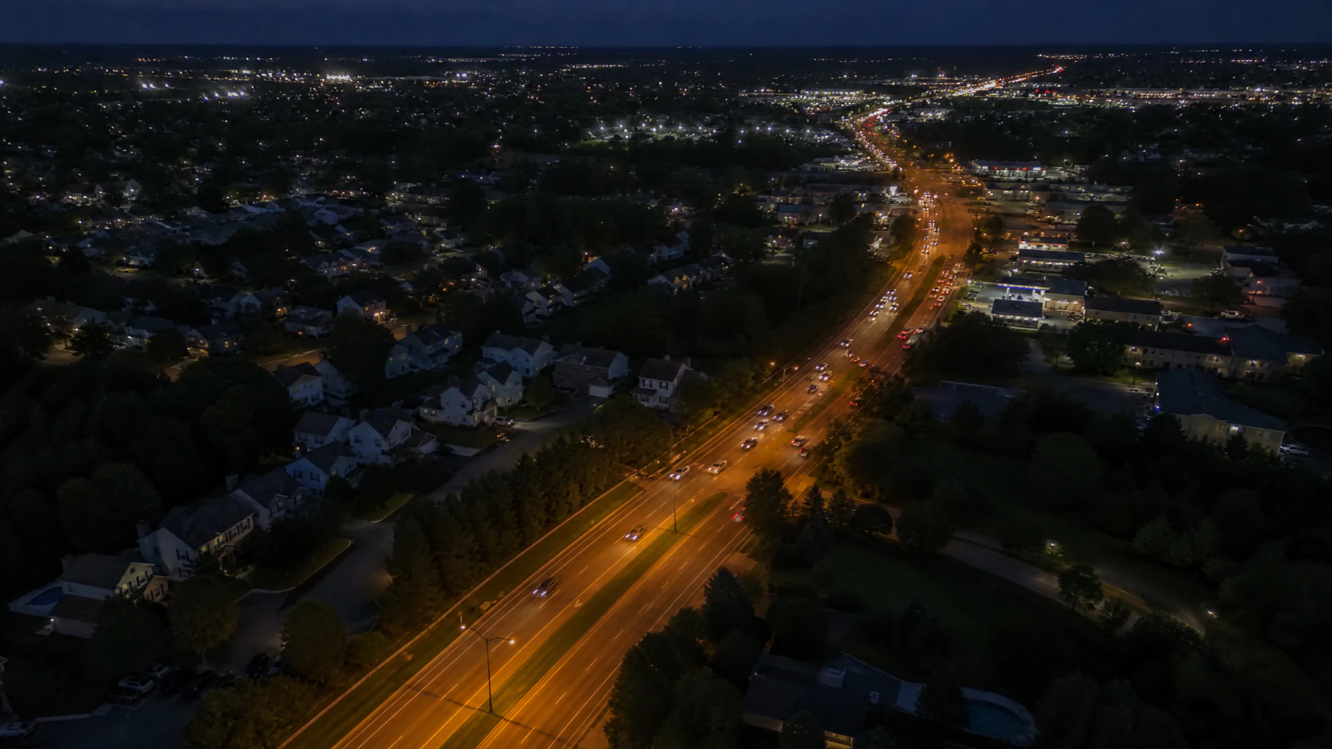 Aerial night view of a multi-lane suburban road glowing orange under streetlights, flanked by residential neighborhoods and commercial properties.