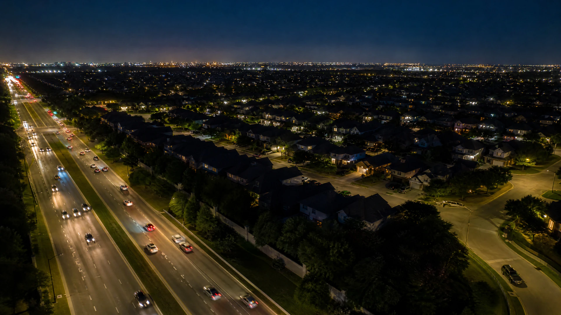 Aerial night view of a multi-lane road with light trails from moving vehicles running alongside a sprawling suburban neighborhood.