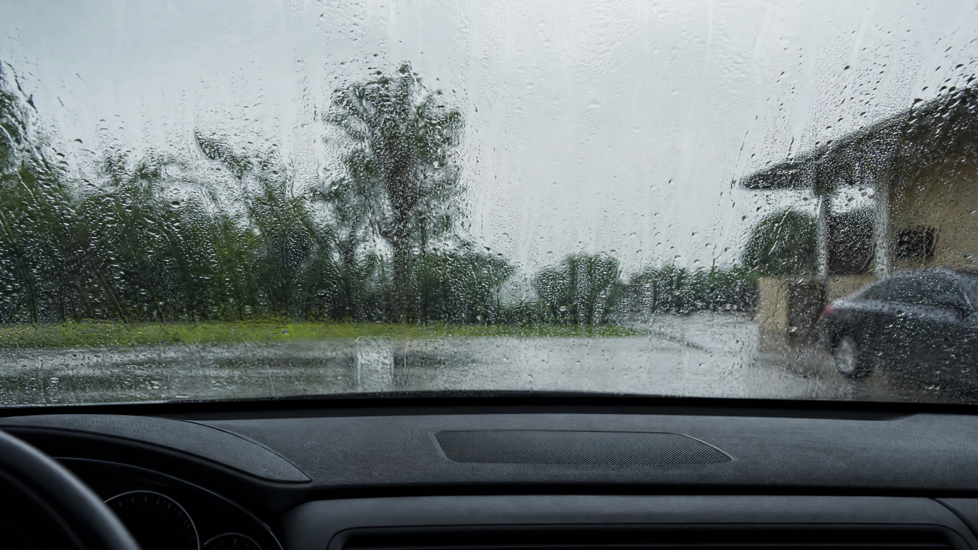 View from a car's interior through a rain-droplet-covered windshield, showing a wet parking area, trees, a building, and another parked car.