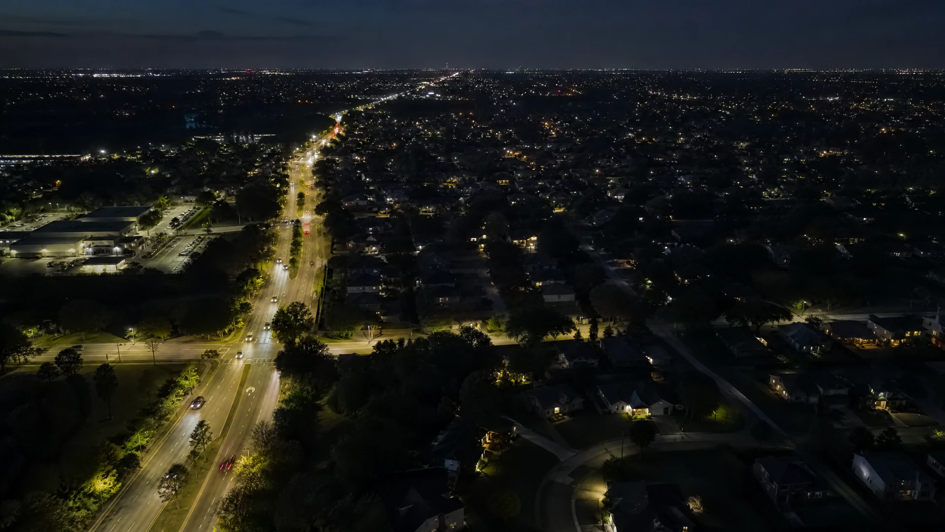 Aerial nighttime view of a suburban landscape divided by a brightly lit multi-lane road, with low-density residential neighborhoods stretching to the horizon.