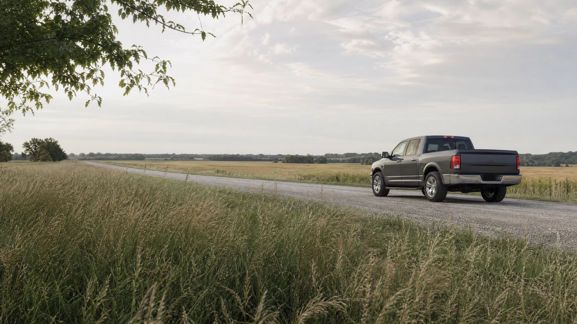 A dark gray pickup truck drives away from the camera on a straight gravel road through open farmland under a partly cloudy sky.