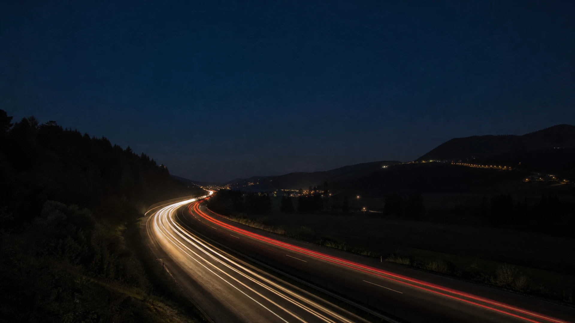 Long-exposure photograph of a curved highway at night, showing white and red light trails left by passing vehicles against a dark landscape and deep blue sky.