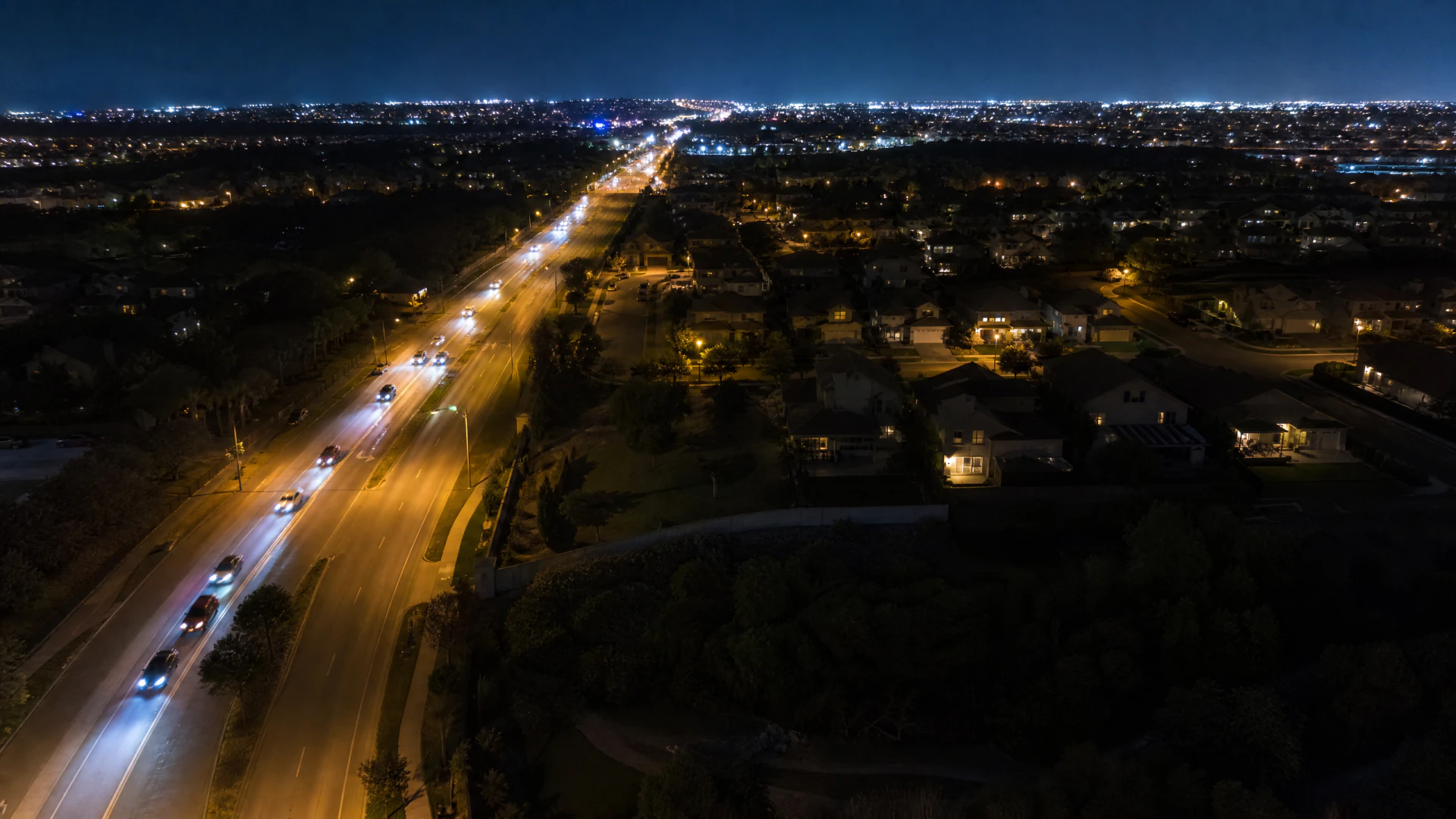 Aerial night view of a multi-lane suburban road lined with street lamps, surrounded by residential neighborhoods extending to a brightly lit city skyline on the horizon.