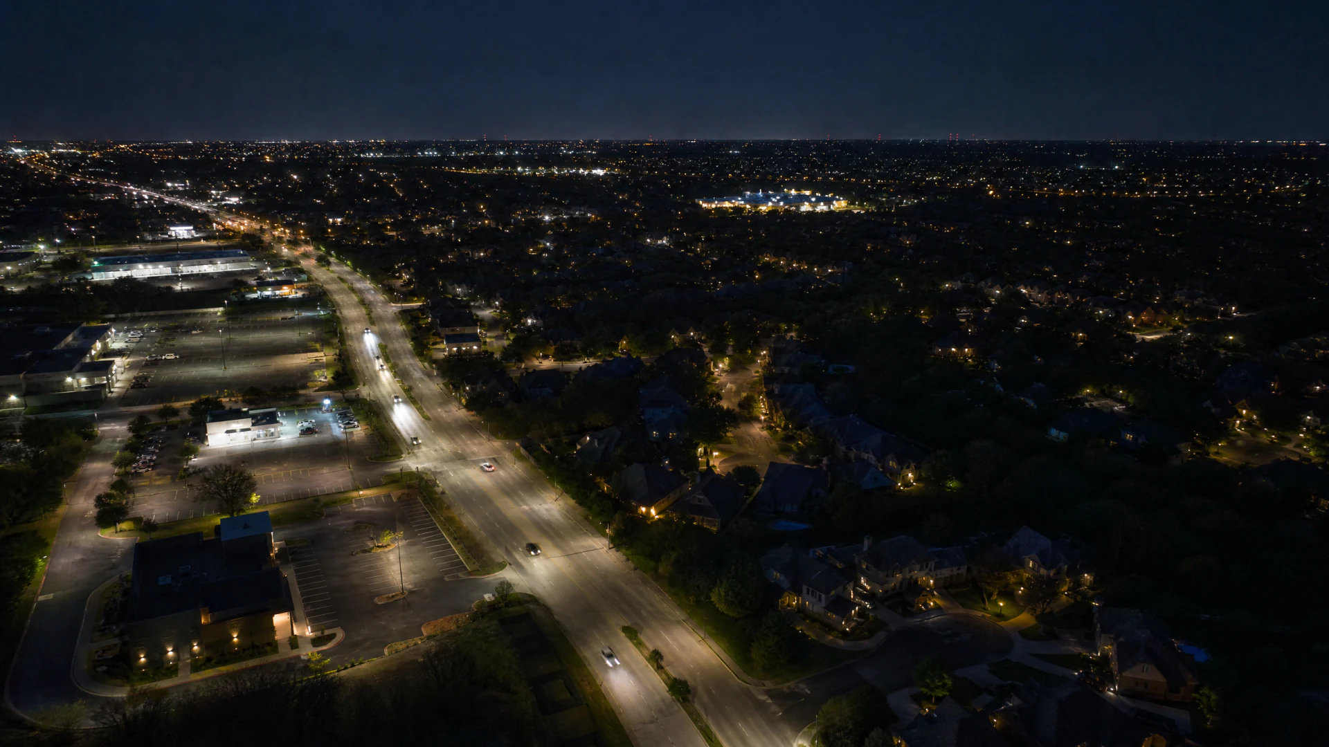 Aerial nighttime view of a suburban road lined with streetlights, bordered by commercial lots and tree-lined residential neighborhoods.