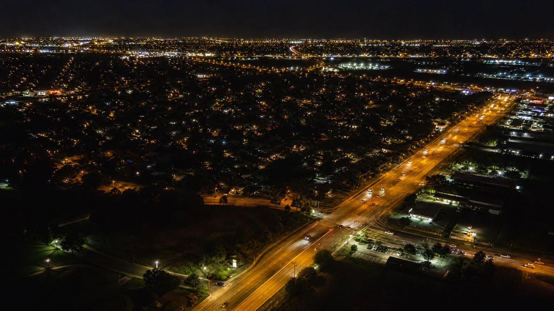 Aerial night view of a suburban city with a lit road intersection surrounded by residential neighborhoods and commercial buildings.