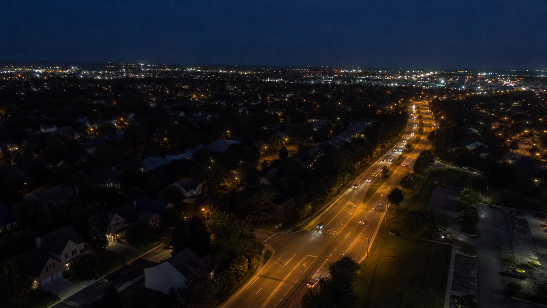 Aerial night view of a illuminated suburban boulevard flanked by residential neighborhoods and trees, with a glowing city skyline in the distance.