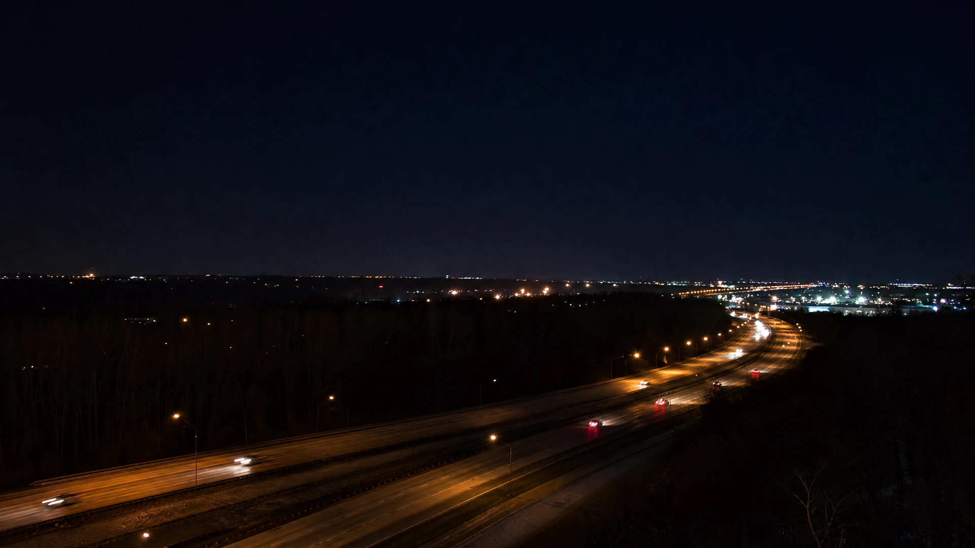 An elevated nighttime view of a lit multi-lane highway curving through a dark, tree-lined landscape with distant city lights on the horizon.