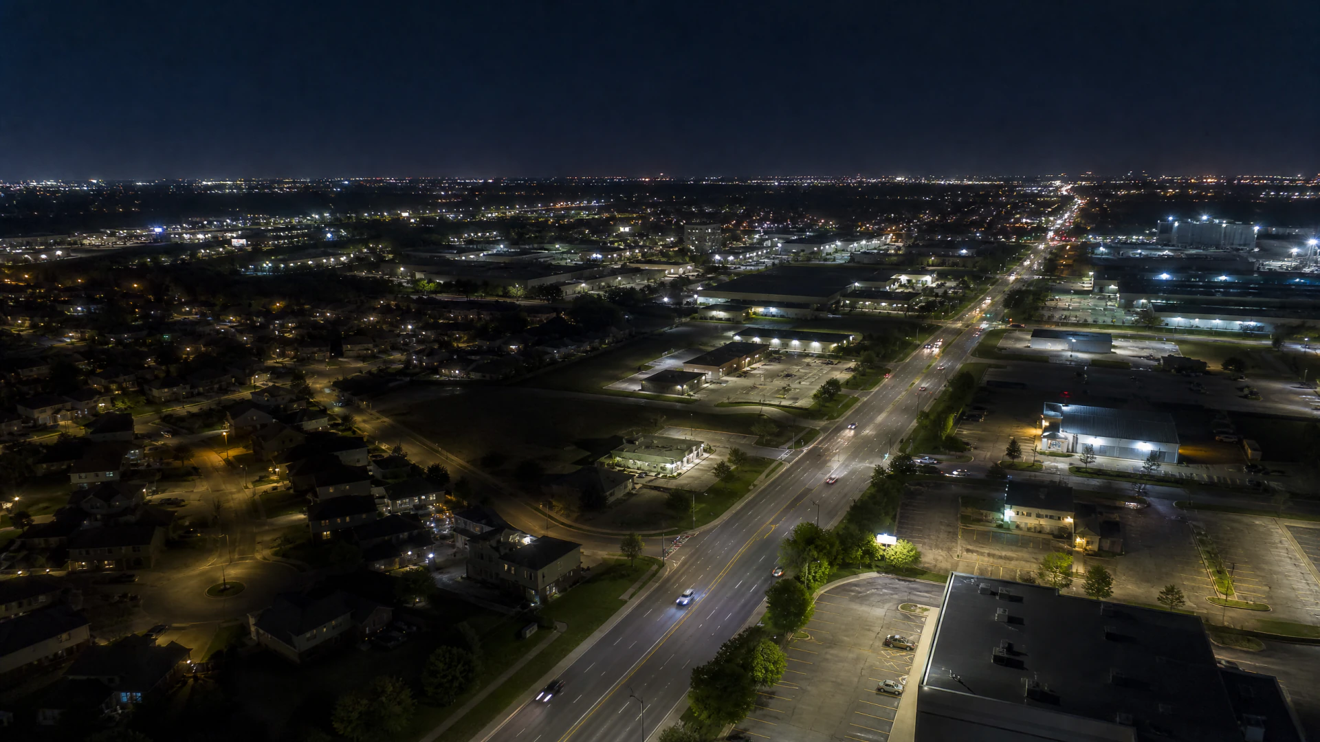 Aerial night view of a wide urban arterial road flanked by commercial properties, parking lots, and residential neighborhoods extending to the horizon.