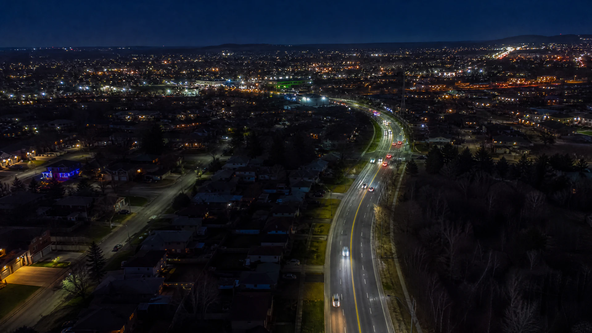 Aerial night view of a curved suburban highway with vehicle light trails, surrounded by a lit residential cityscape and dark tree lines.