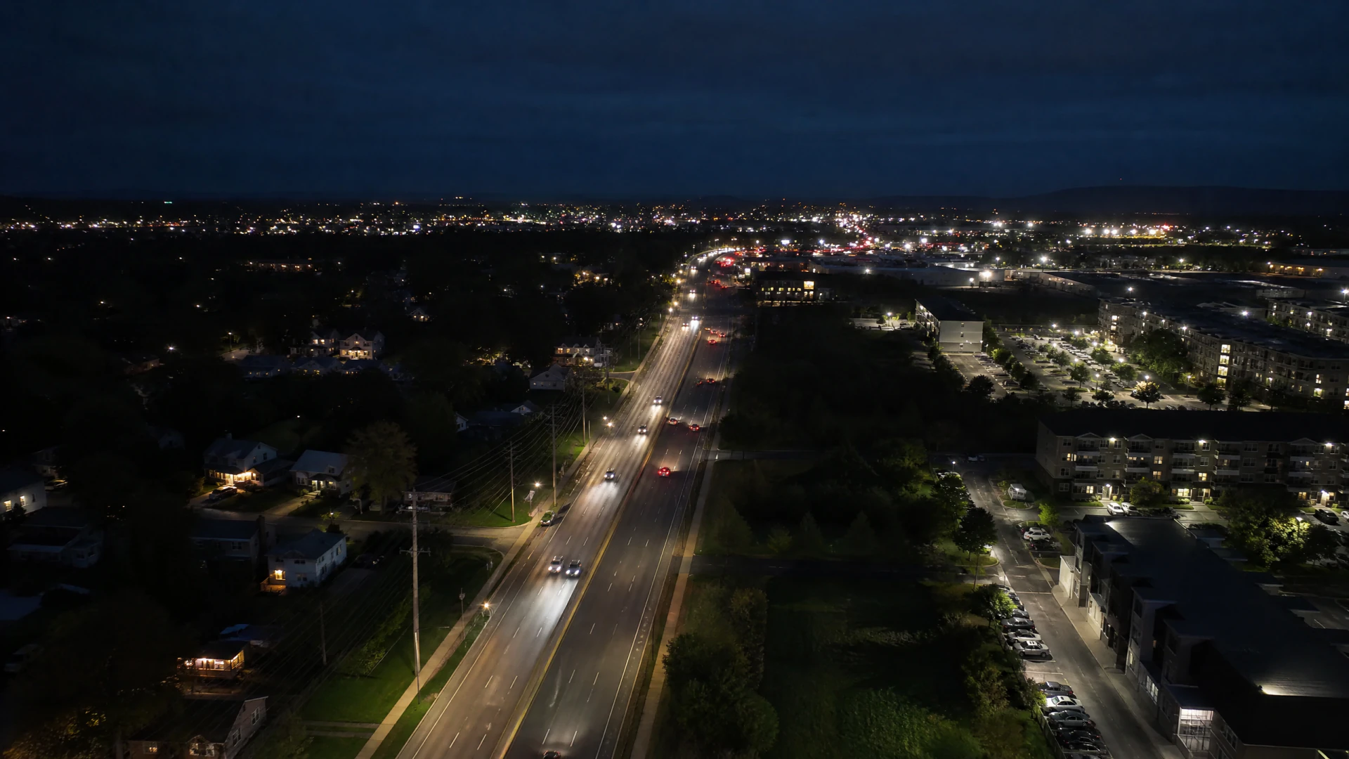 Aerial night view of a suburban divided highway with moving traffic, flanked by lit residential neighborhoods on the left and commercial buildings on the right.