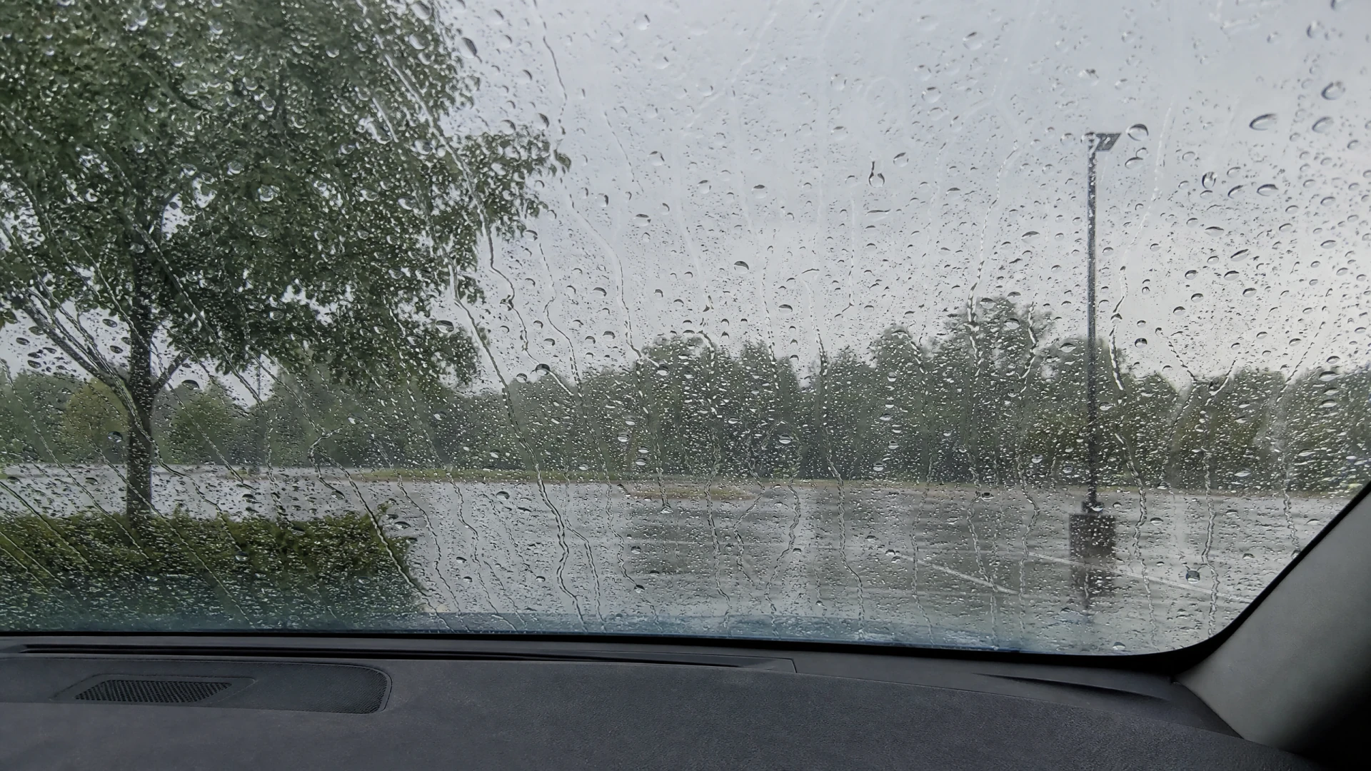 A rain-covered car windshield looking out over a wet parking lot lined with trees under a grey sky.