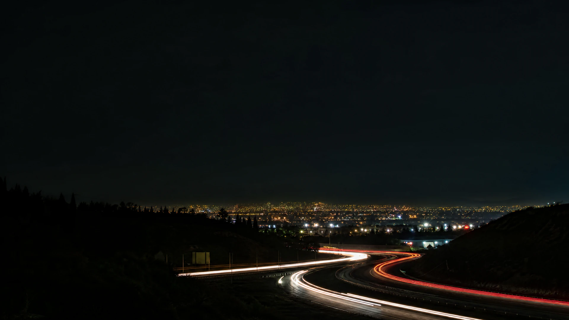Long-exposure photograph of a curved highway at night, showing streaking white and red light trails from vehicles with a illuminated city skyline in the distance.