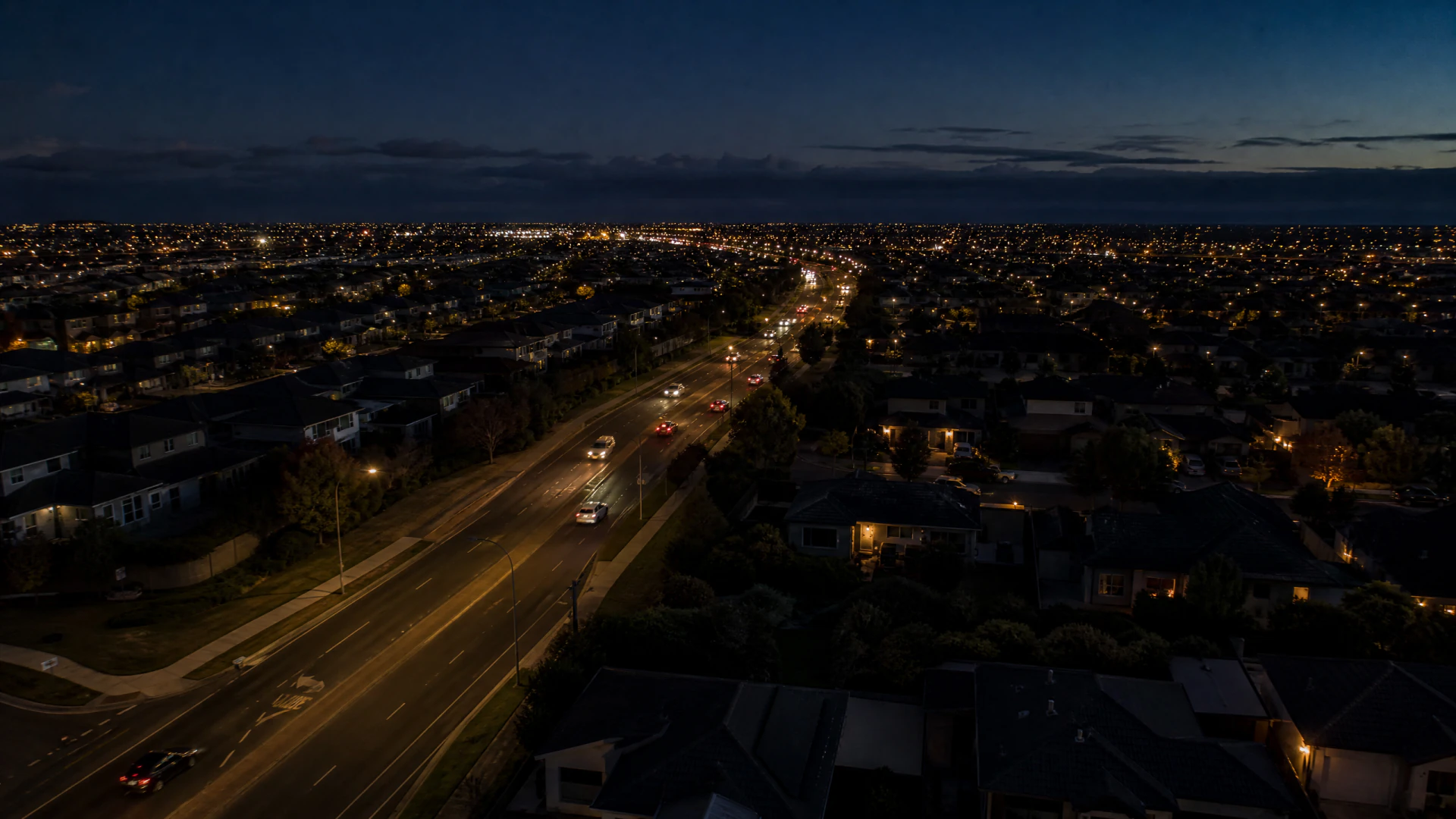 Aerial night photograph of a suburban neighborhood with a wide illuminated road running through the center, surrounded by houses and trees.