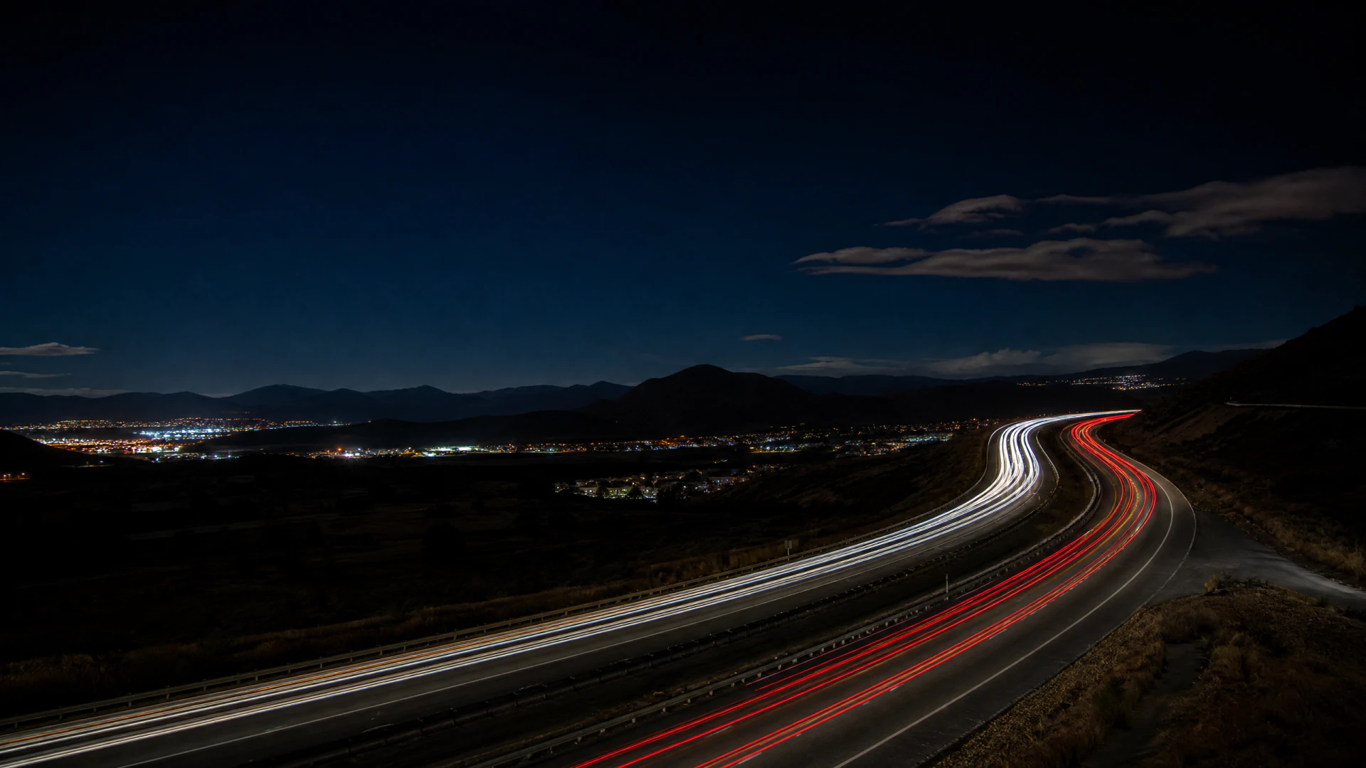 Long-exposure photograph of a highway at night showing streaked white and red light trails curving toward a illuminated valley below.
