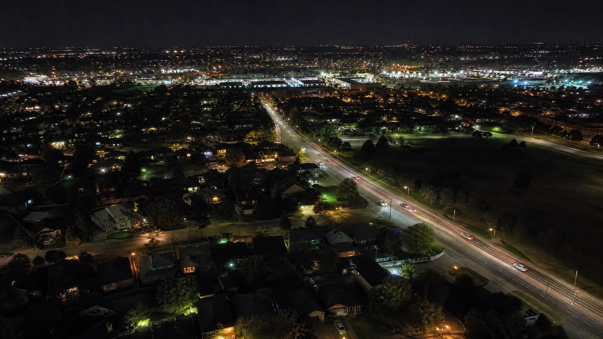 Aerial nighttime view of a suburban neighborhood divided by a multi-lane road with vehicle light trails and a lit commercial district in the distance.