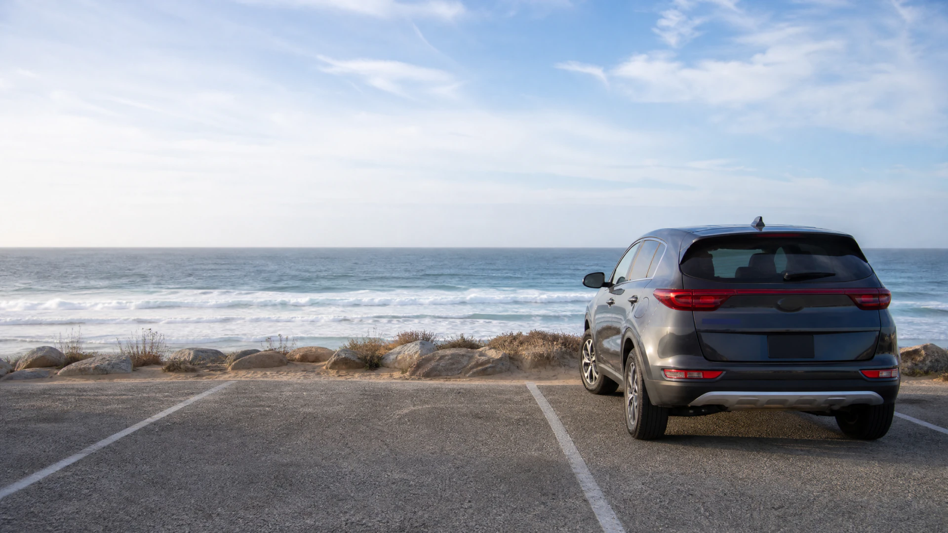 A dark gray SUV parked in a beachside lot, viewed from the rear, with ocean waves and a rocky shoreline in the background.