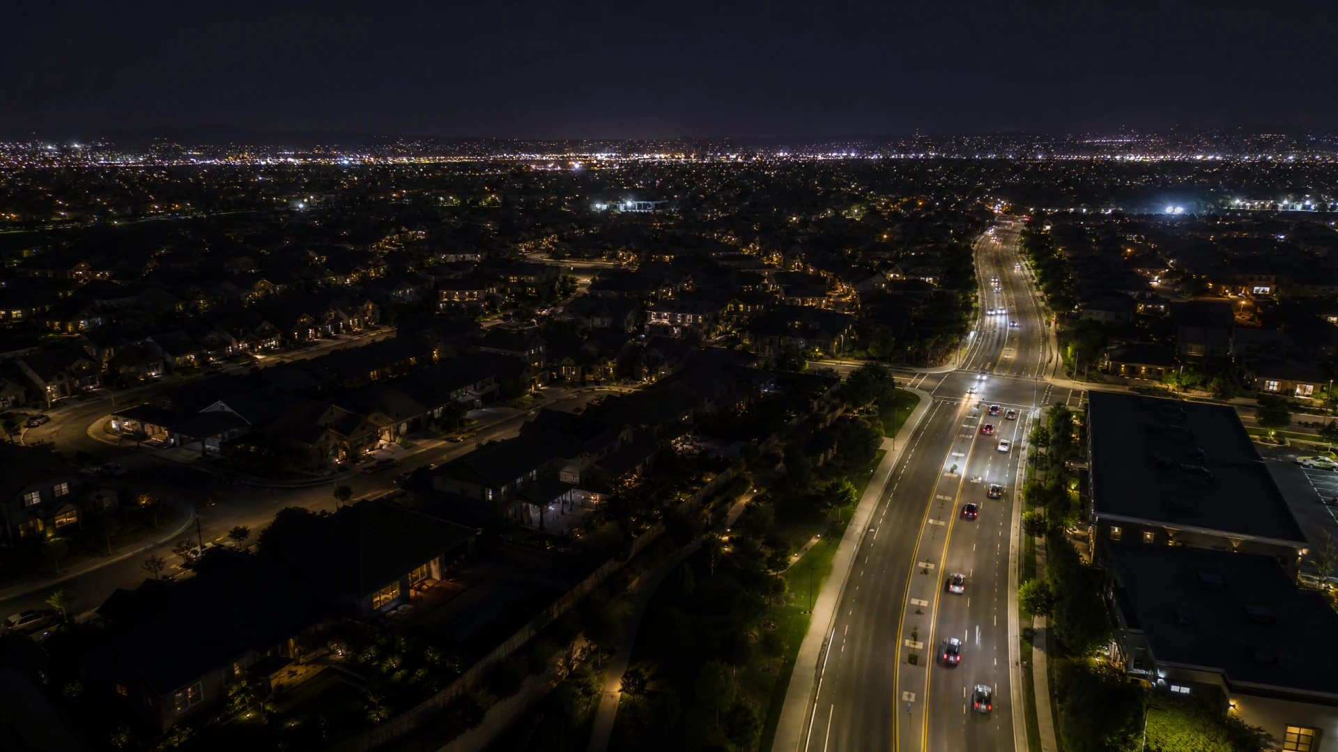 Aerial night photograph of a wide multi-lane road cutting through a sprawling suburban landscape illuminated by streetlights and city glow.