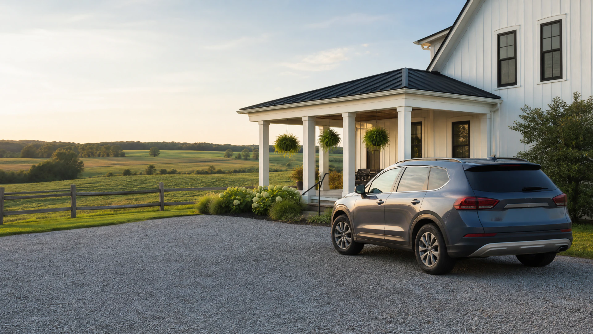 A gray SUV parked on a gravel driveway in front of a white farmhouse with a covered porch and open countryside beyond.