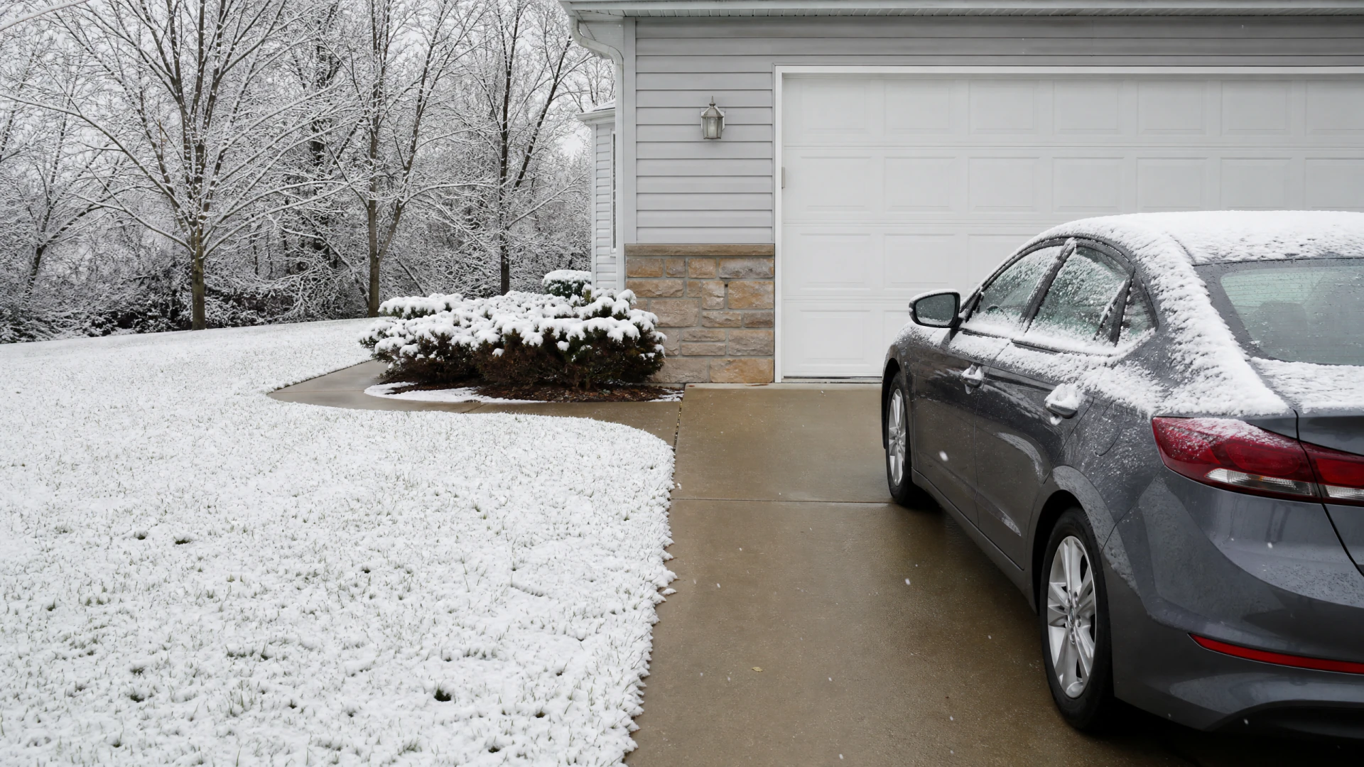 A snow-covered gray sedan parked in the driveway of a house with a white garage door, surrounded by snow-dusted trees and shrubs.
