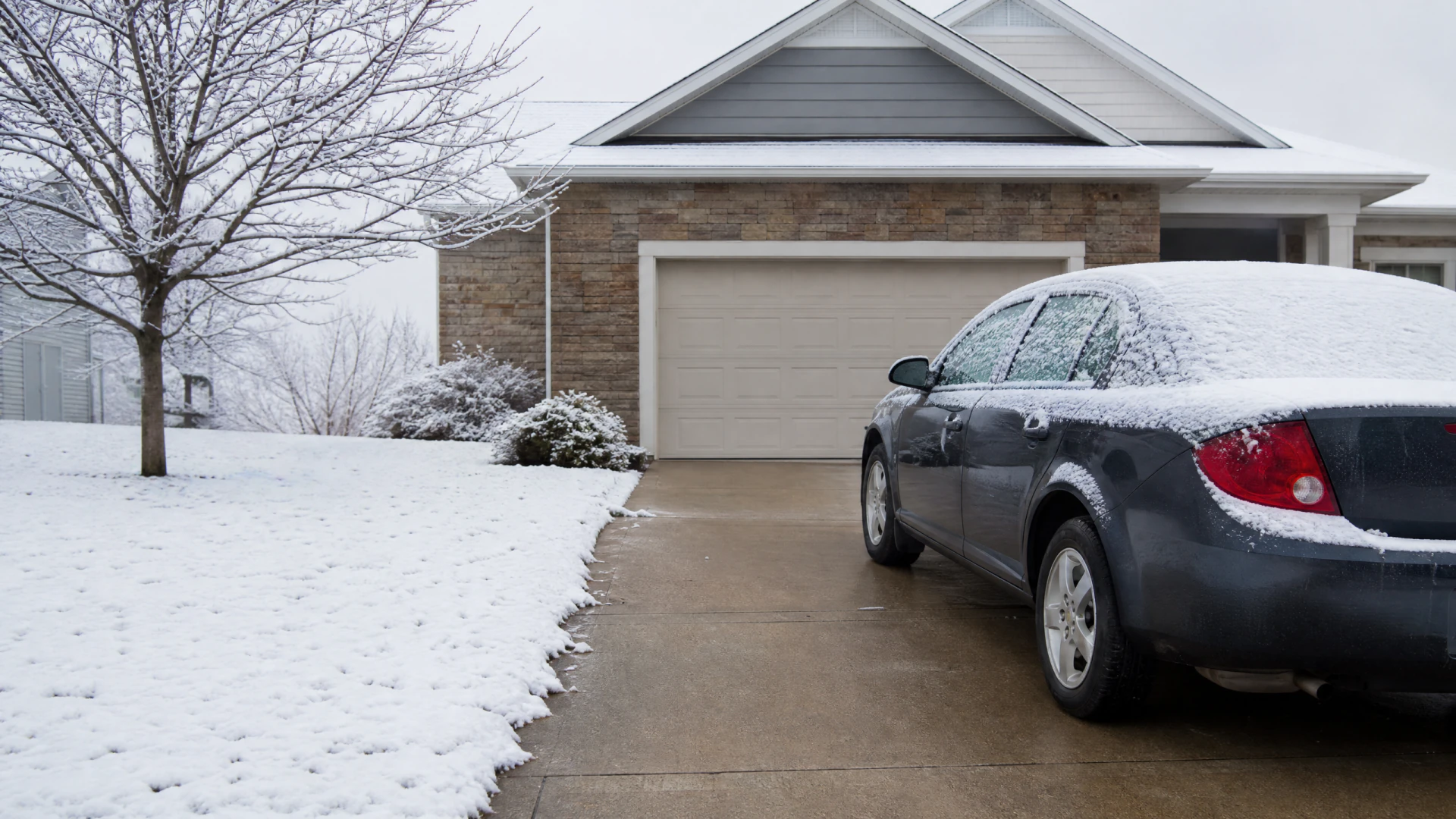 A snow-dusted dark blue sedan parked in a concrete driveway in front of a two-car garage on a winter day.