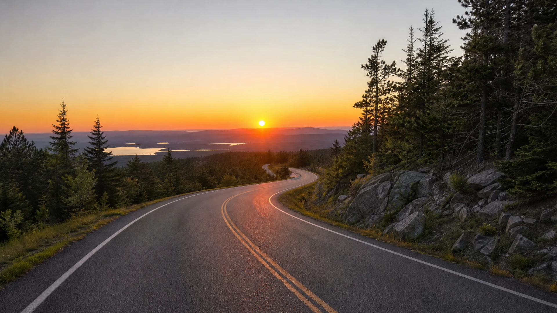 A winding two-lane road curves through a forested mountain landscape at sunset, with rocky outcrops on one side and a lake visible in the distant valley below.
