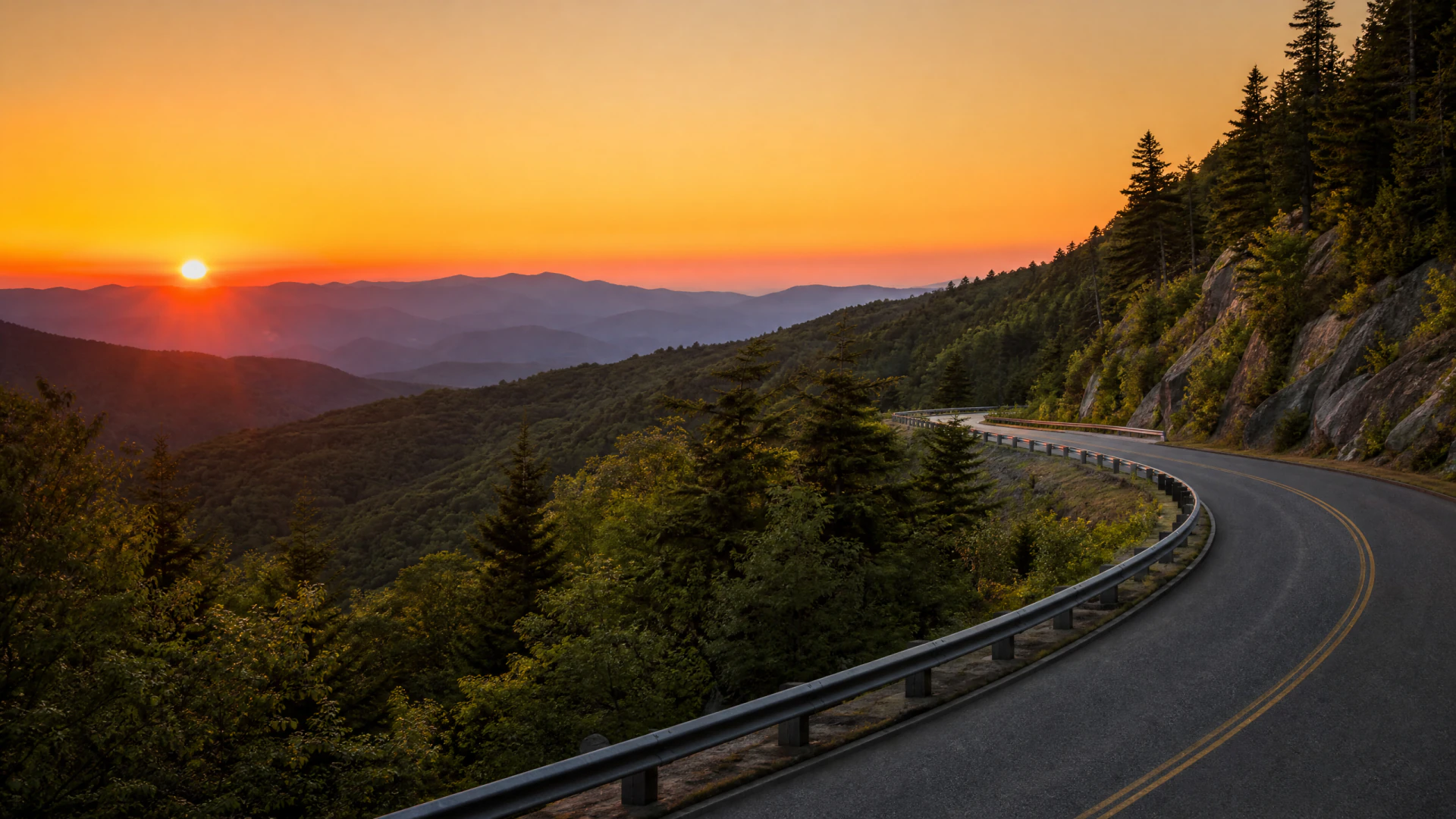 A winding mountain road with a metal guardrail curves along a forested hillside at sunset, with multiple hazy mountain ridges visible in the background.