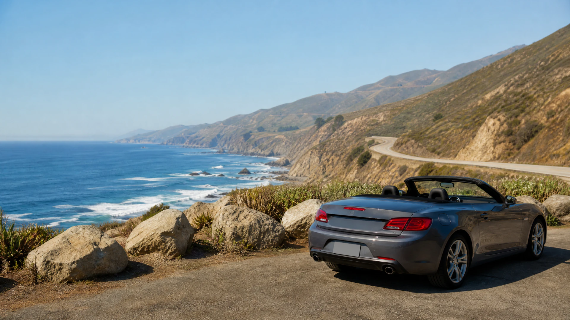 A silver convertible with its roof down is parked at a scenic coastal overlook, with rocky cliffs, a winding highway, and the ocean visible in the background.