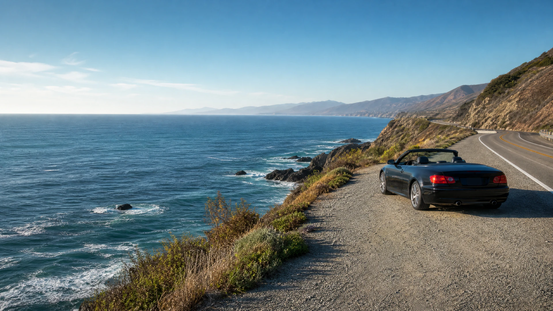 A black convertible car parked on a gravel shoulder beside a two-lane coastal highway overlooking rocky cliffs and open ocean.