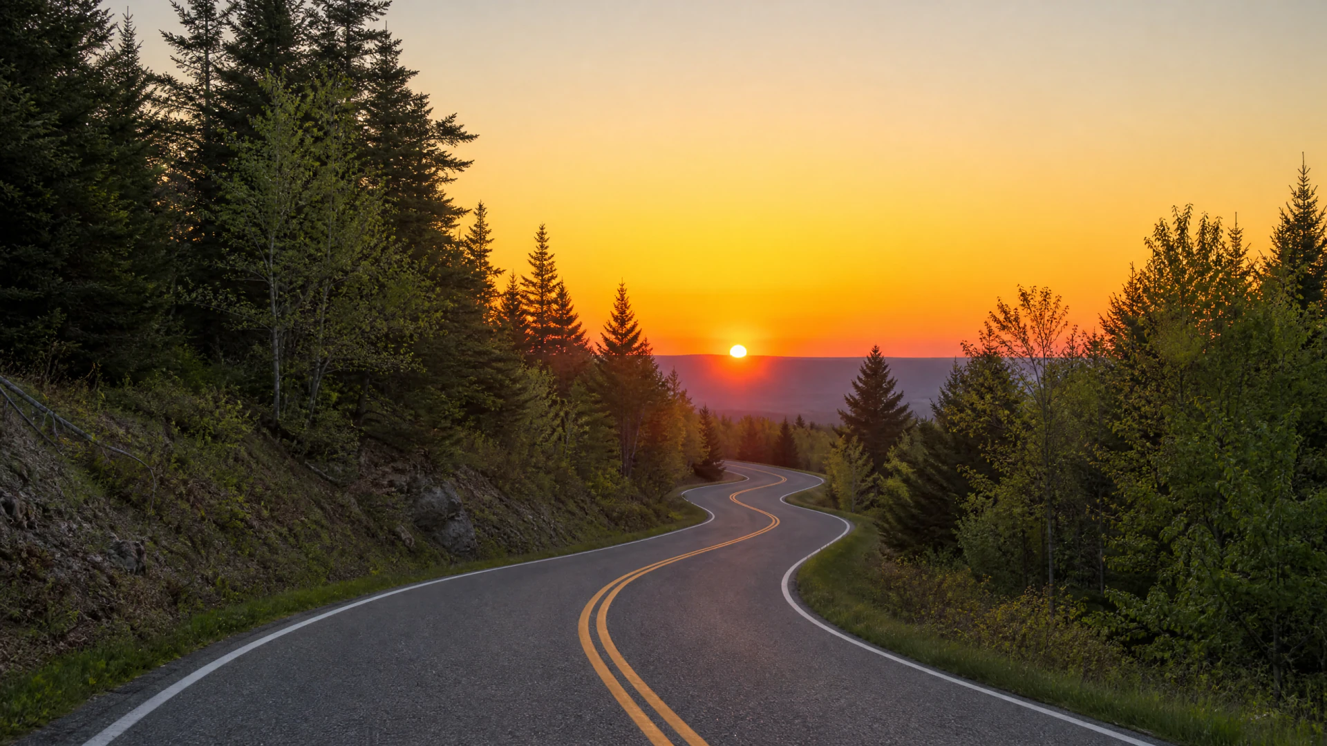 A winding two-lane road curves through a forested landscape beneath a bright orange sunset sky.
