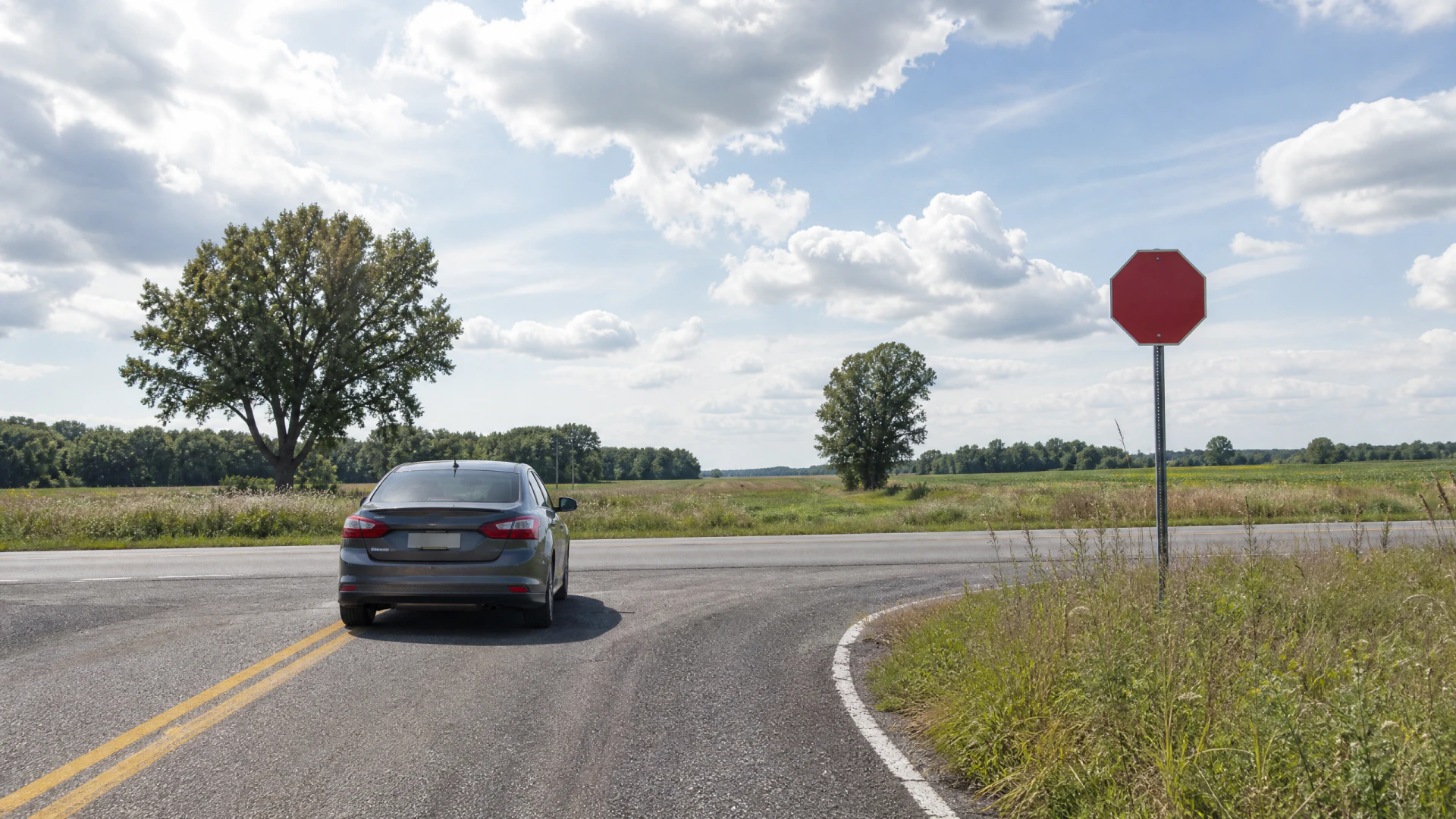 A gray sedan pauses at a rural road intersection next to a blank red stop sign.
