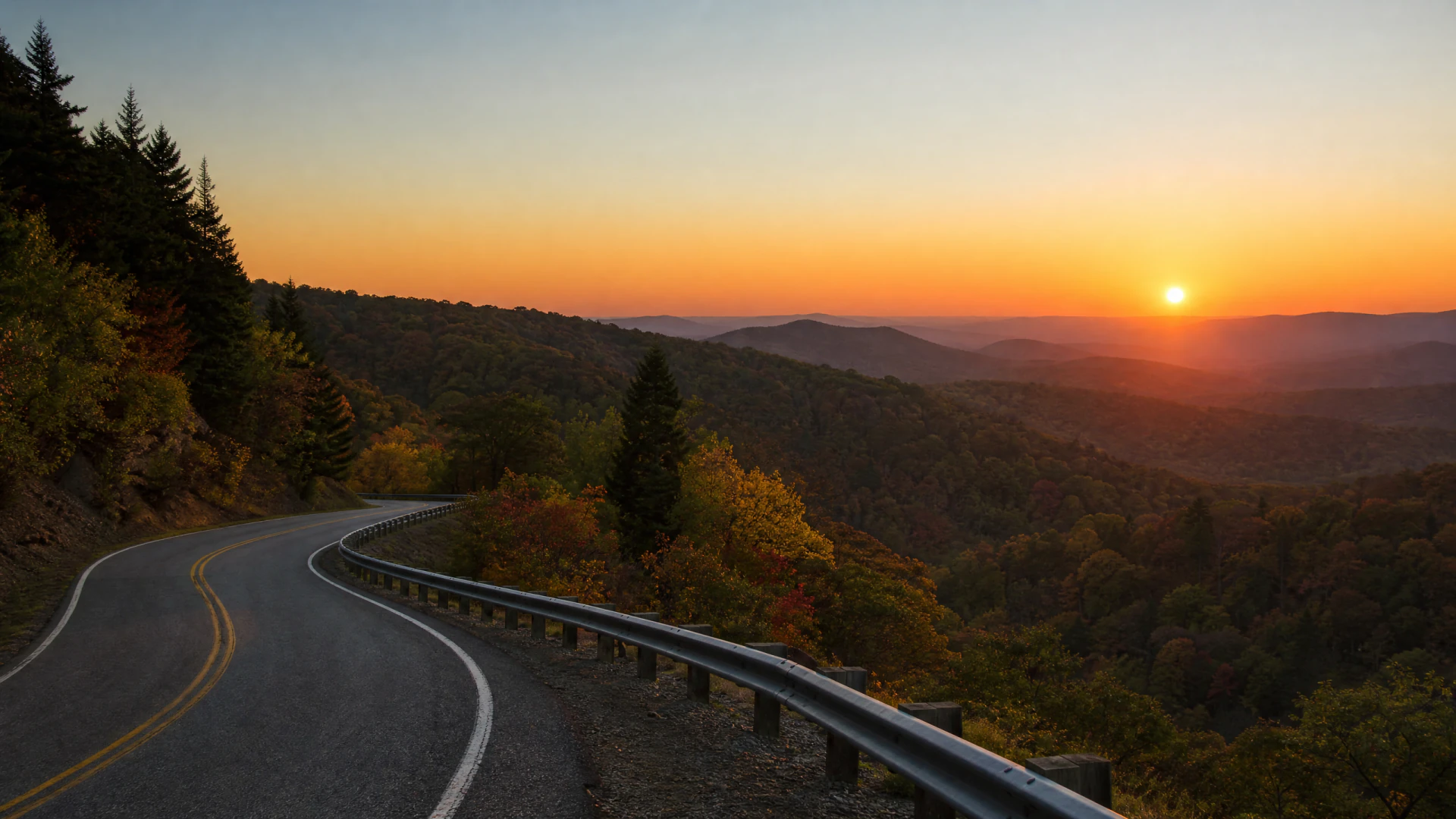 A winding two-lane mountain road curves past a metal guardrail at sunset, overlooking a vast forested valley with rolling hills.