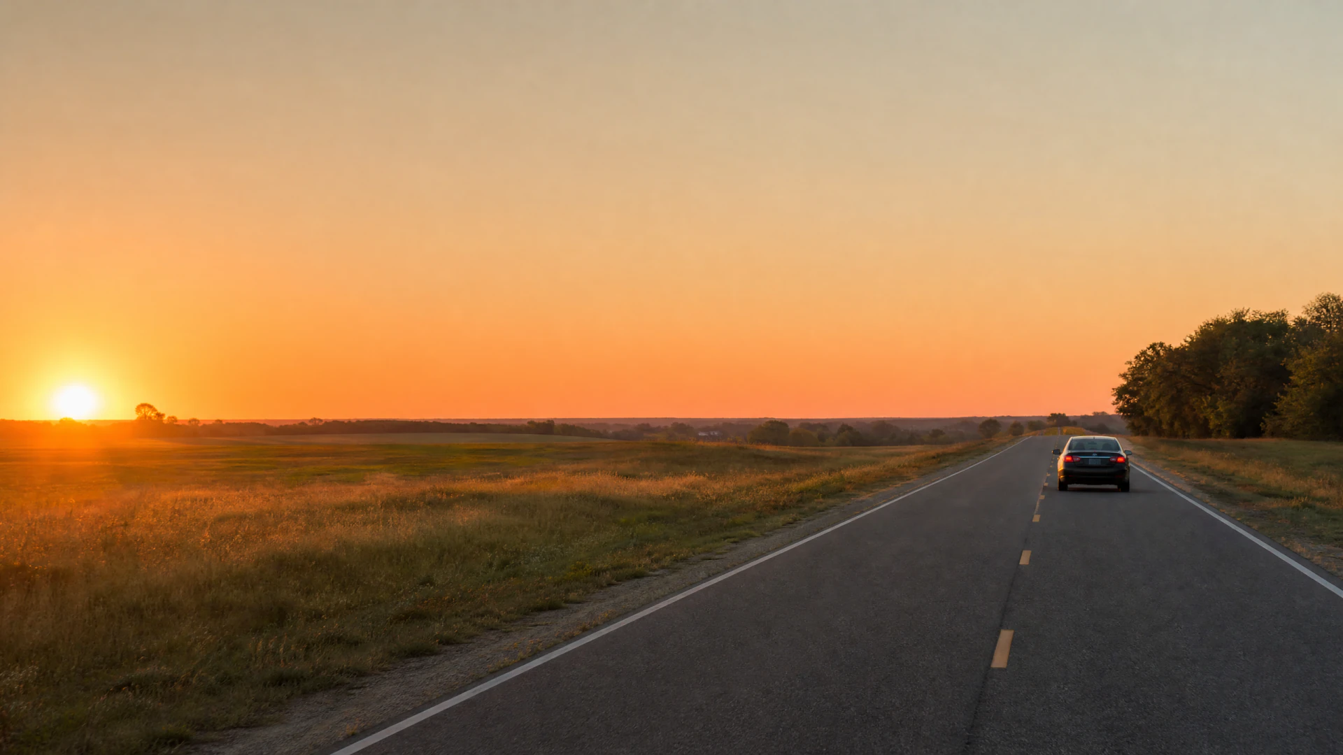 A car drives away from the camera on a straight rural highway through open fields at sunset.