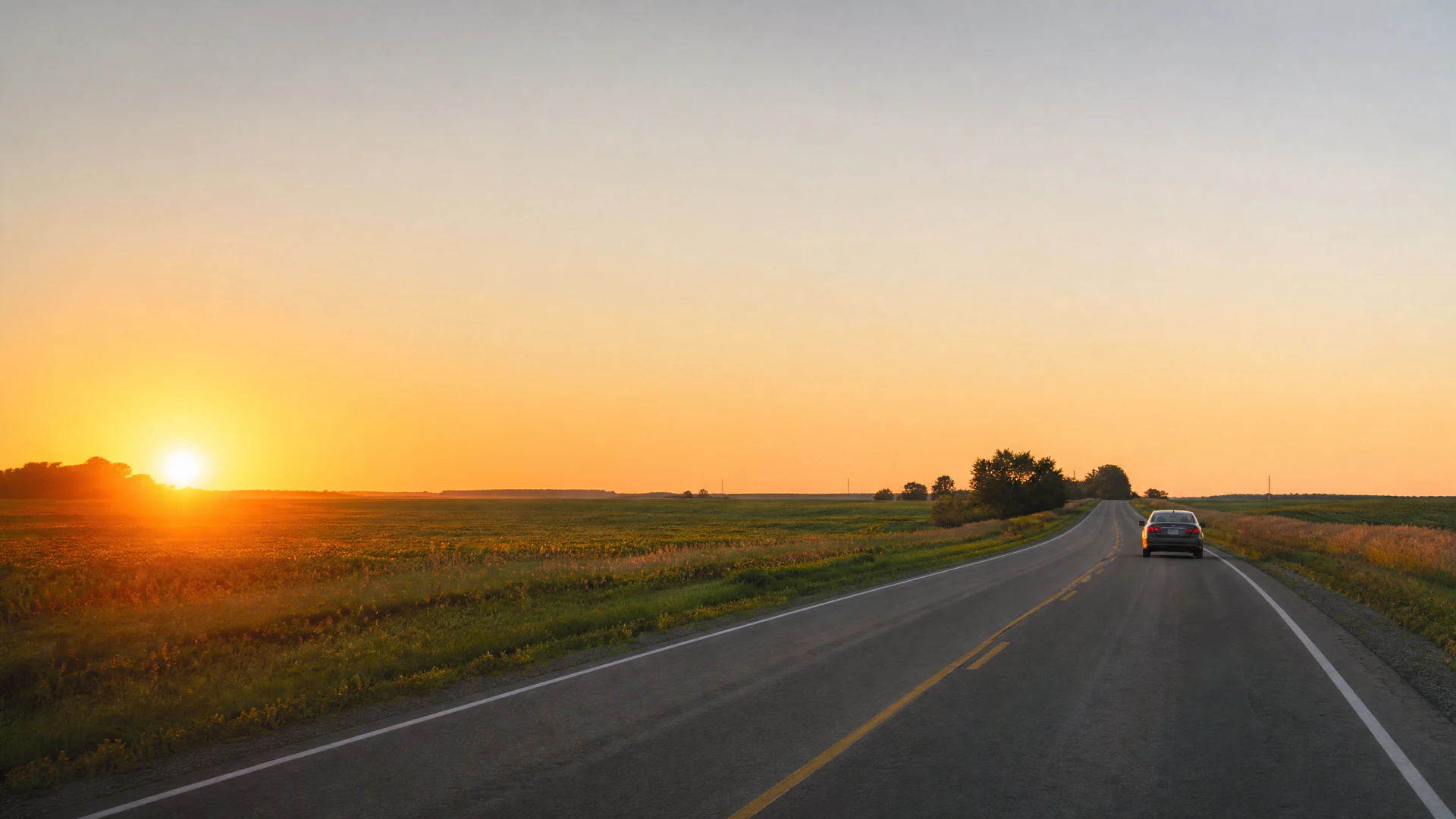 A car drives away from the camera on a two-lane rural road at sunset, with flat agricultural fields stretching to the horizon on both sides.