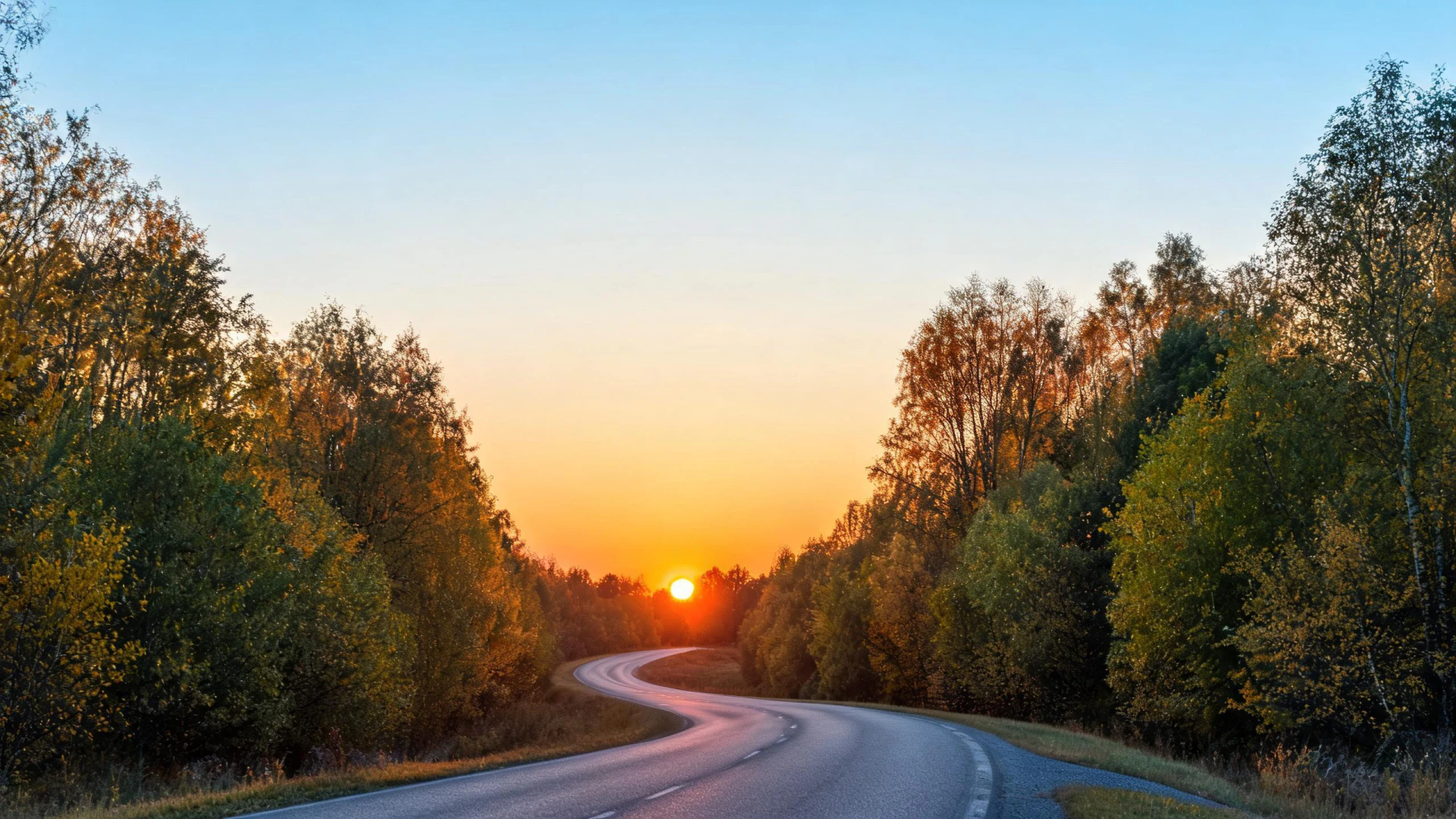 A winding two-lane road flanked by autumn trees leads toward a low sun setting on the horizon.