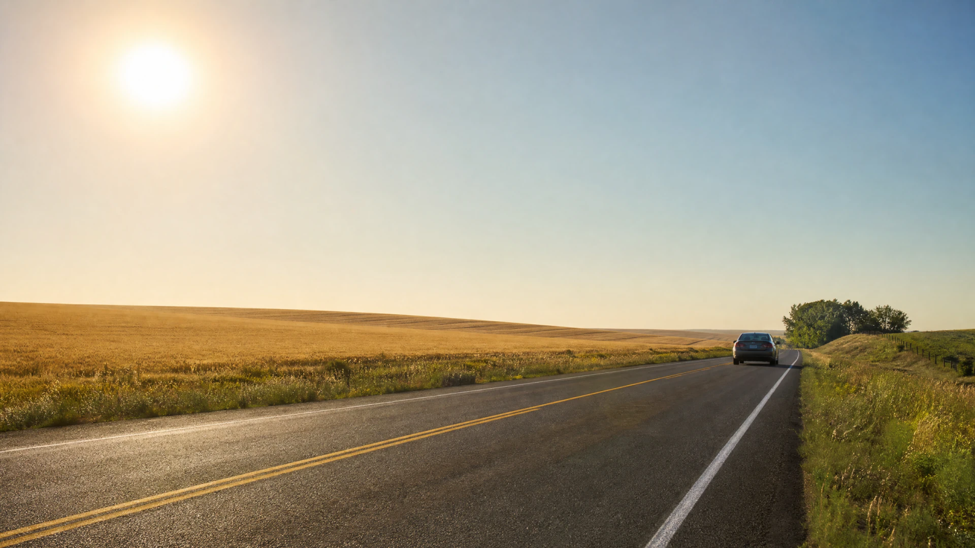 A sedan travels down a straight two-lane rural highway bordered by golden wheat fields and a clear blue sky.
