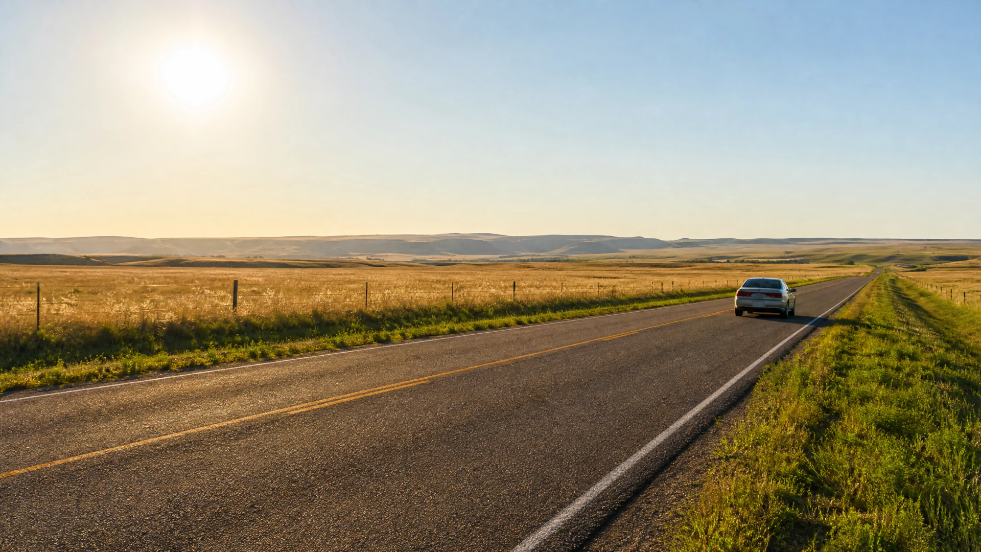 A silver sedan travels down a straight rural highway flanked by golden prairie grassland and distant hills.