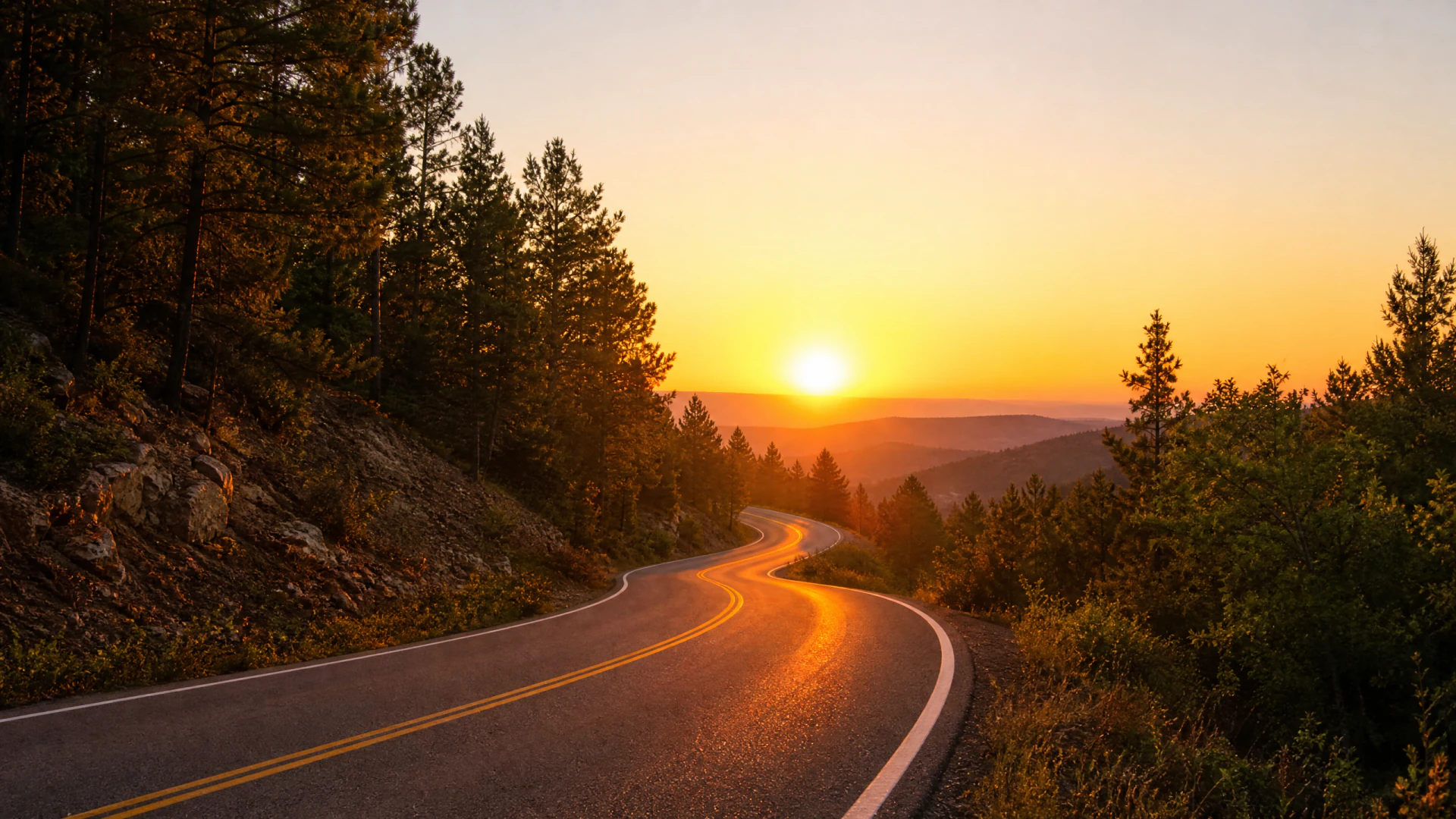 A winding two-lane mountain road curves through a pine forest toward a bright orange sunset over distant hills.