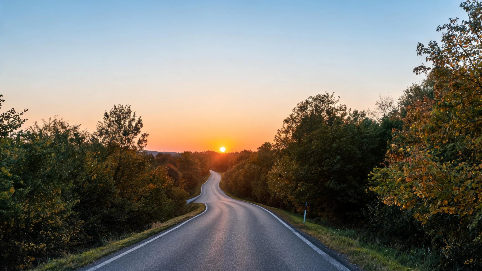 A winding two-lane road flanked by autumn trees leads toward a low sun on the horizon.