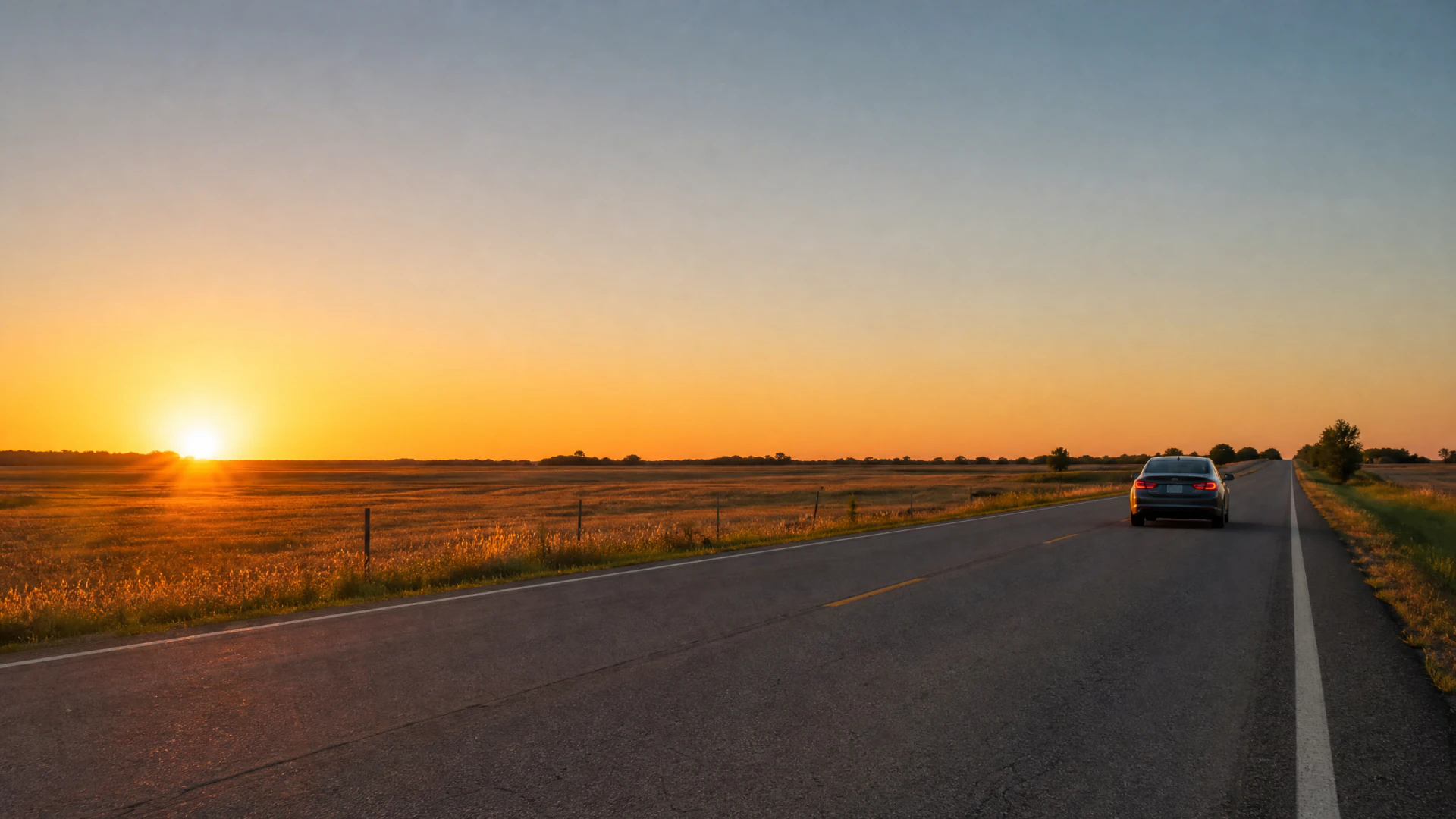 A sedan travels down a straight rural highway at sunset, surrounded by flat grasslands under a clear sky.