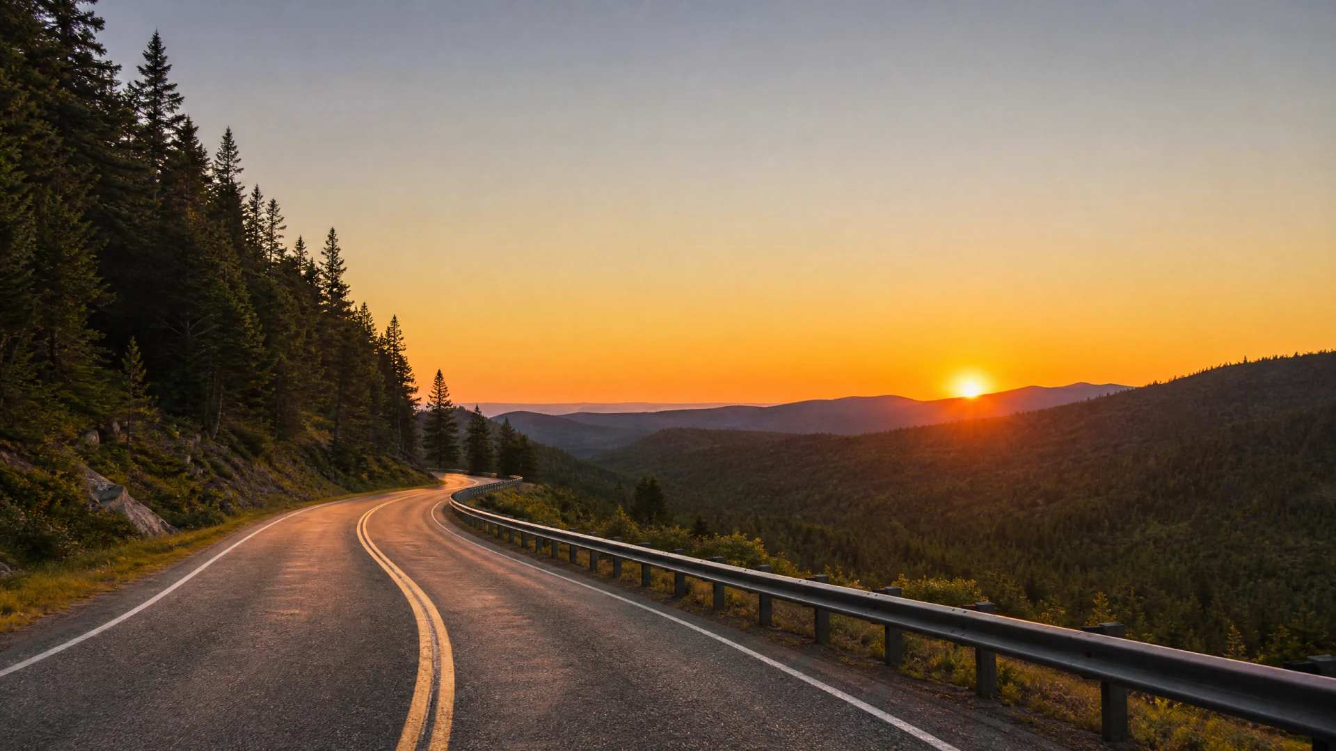 A winding two-lane mountain road bordered by conifer trees and a metal guardrail, photographed at sunset with rolling forested hills in the distance.