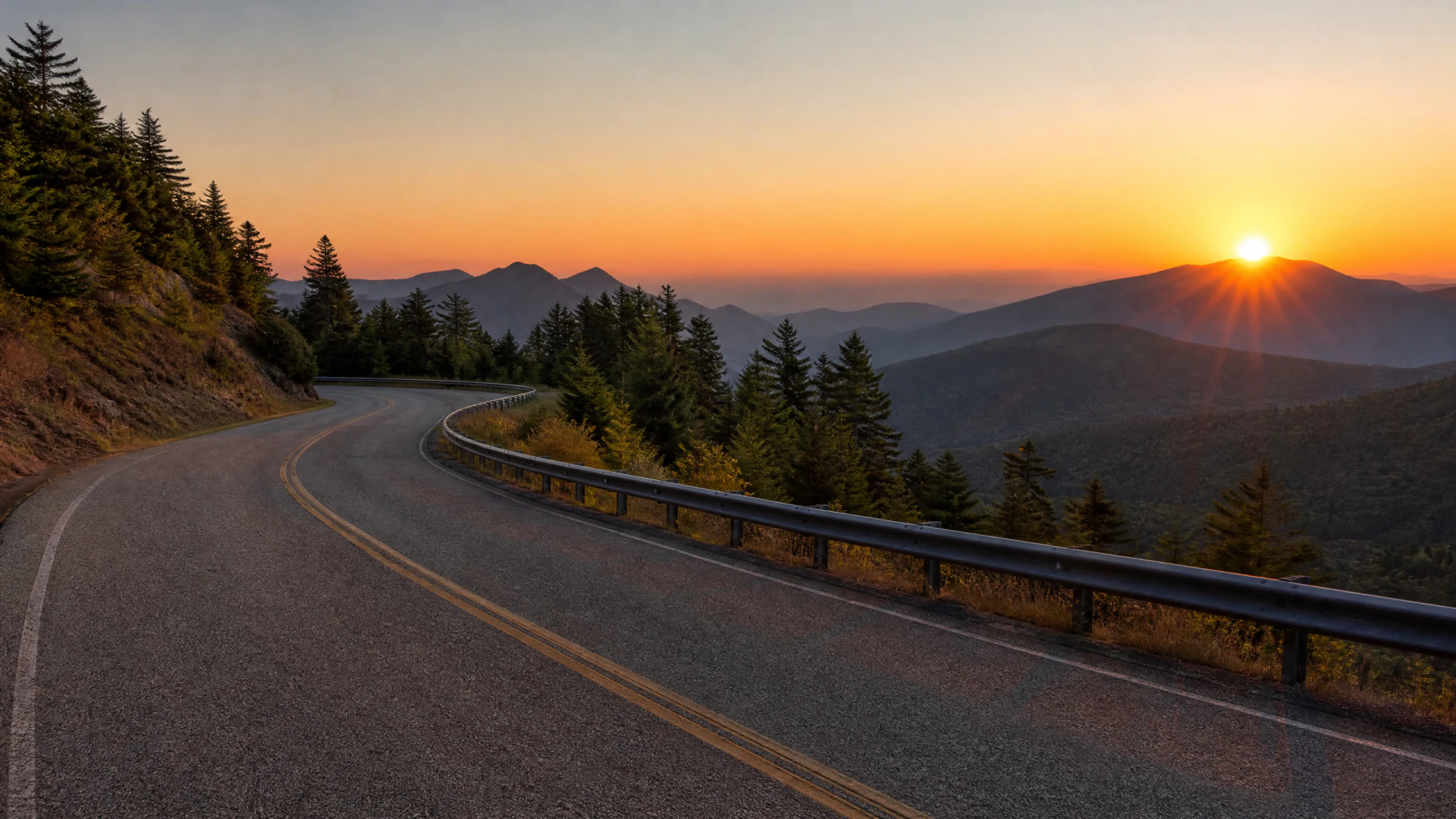 A winding two-lane mountain road curves past a guardrail and conifer trees at sunset, with layered mountain ridges visible in the distance.