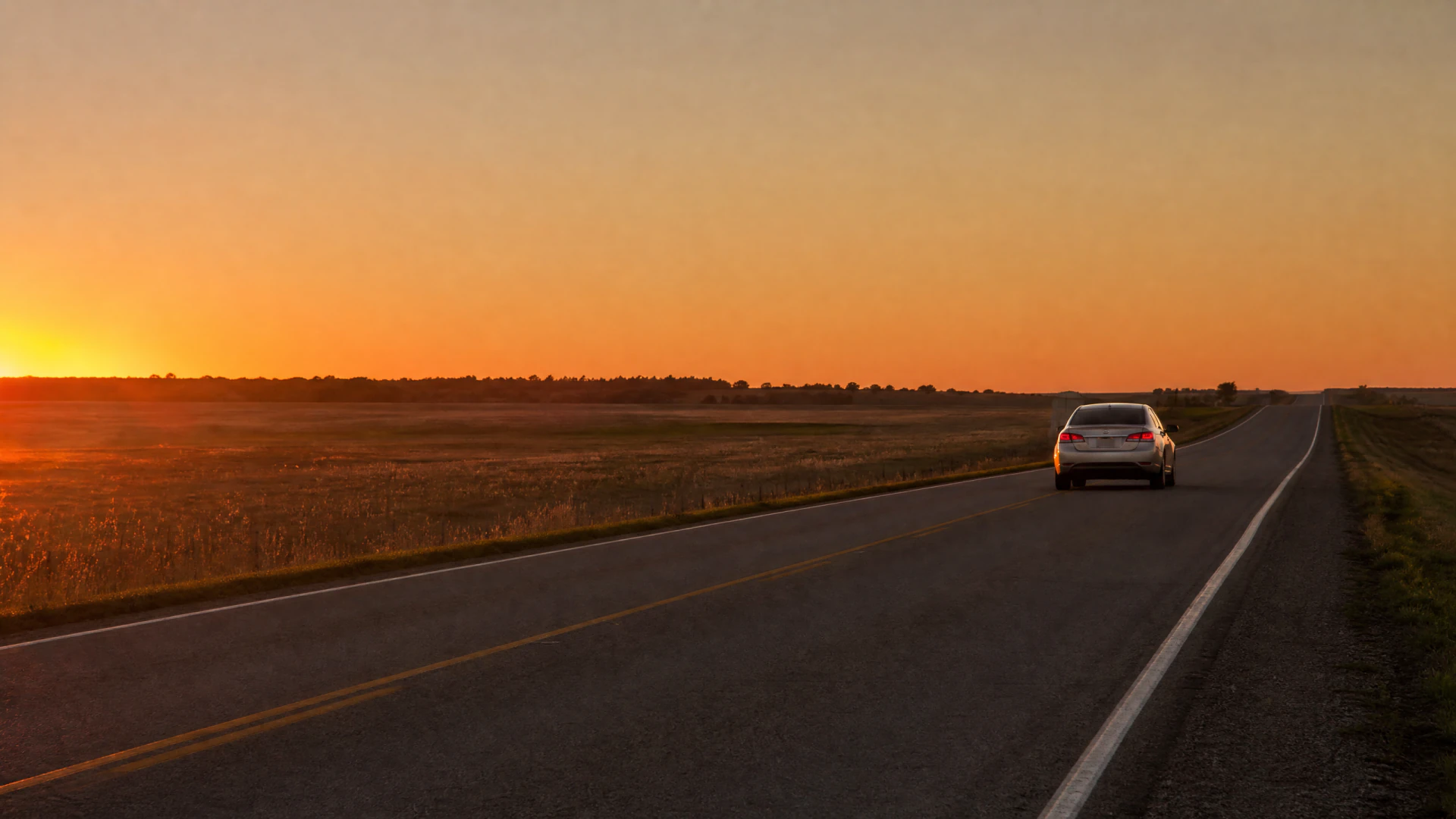 A silver sedan drives along a straight two-lane rural highway at sunset, flanked by flat open fields under an orange sky.