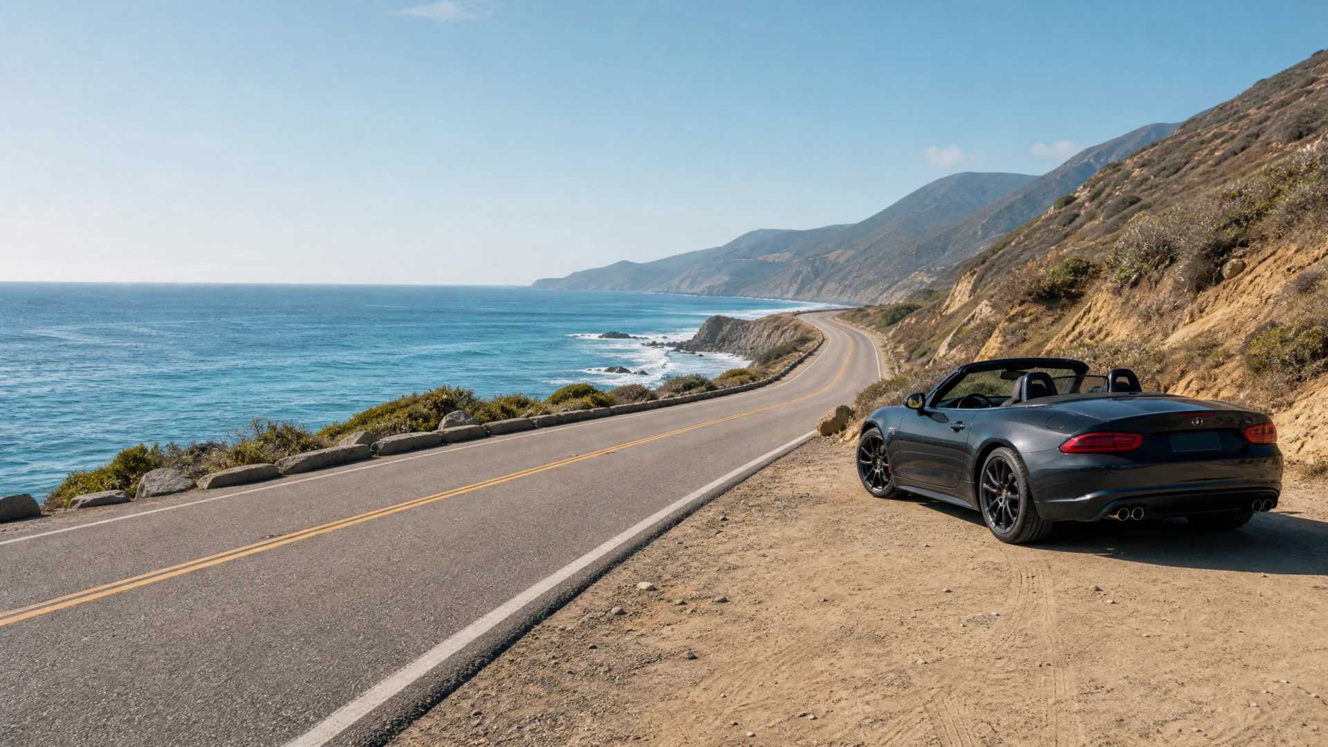 A dark gray open-top sports car parked beside a winding coastal highway with ocean views and hillside terrain.