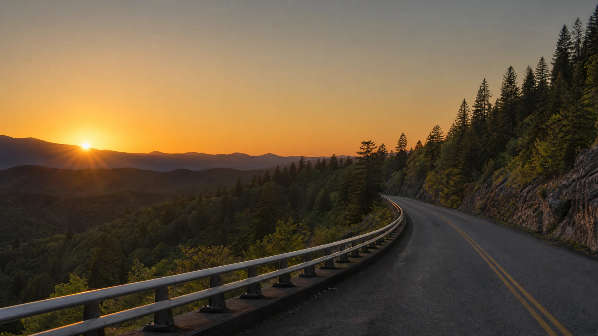 A winding two-lane road curves along a forested mountain hillside at sunset, with a metal guardrail on the left and conifer trees on the right.