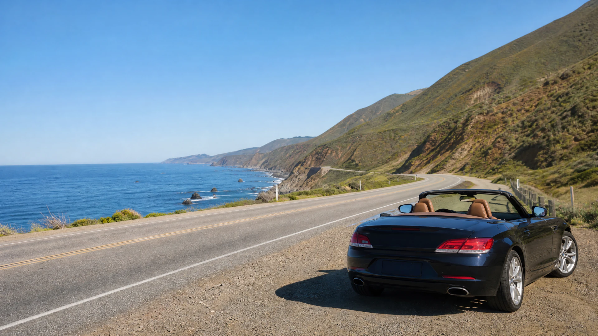 A dark blue open-top convertible parked on the shoulder of a winding coastal highway, with ocean cliffs and hills in the background.