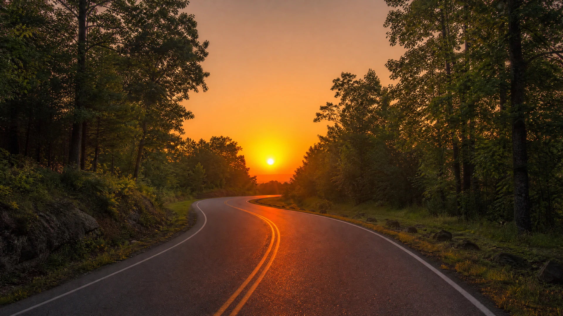 A winding two-lane road lined with green forest trees leads toward a vivid orange sunset on the horizon.