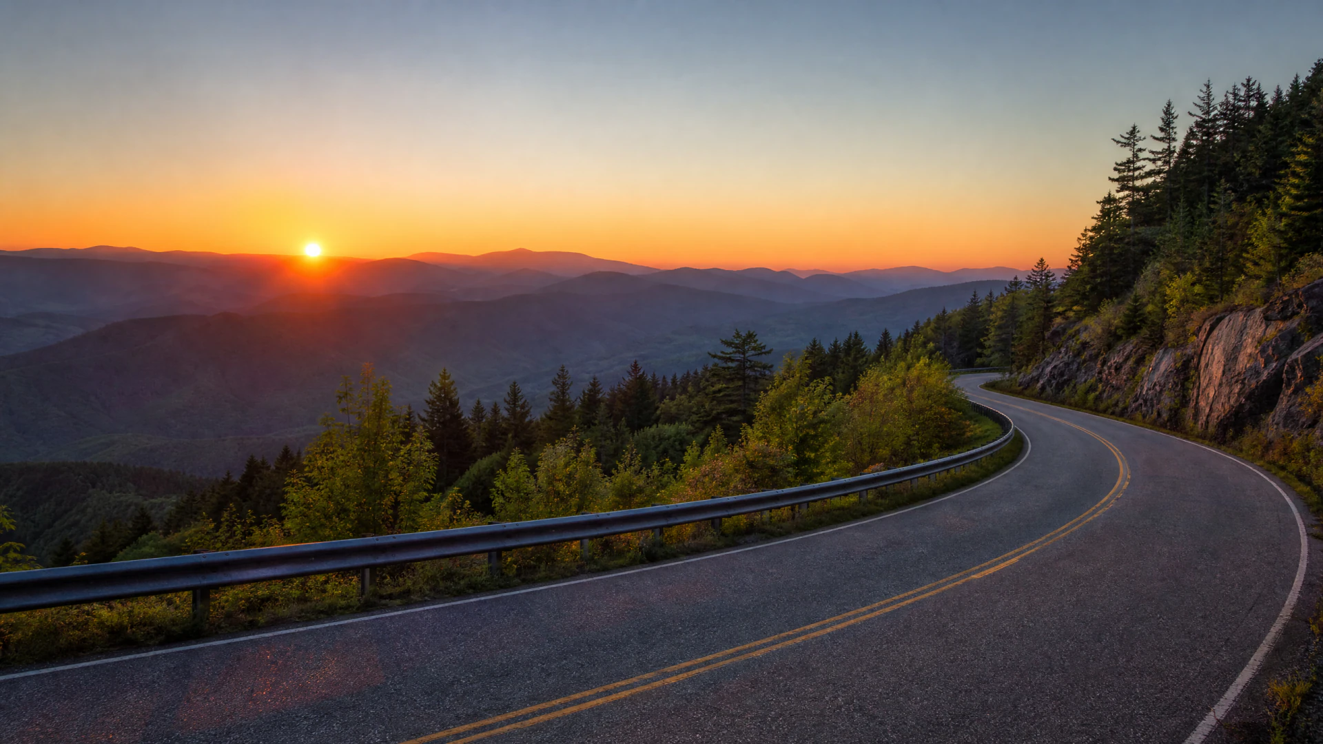 A winding two-lane road curves along a forested mountain ridge at sunset, with a guardrail on the left and rolling mountain peaks glowing orange in the distance.