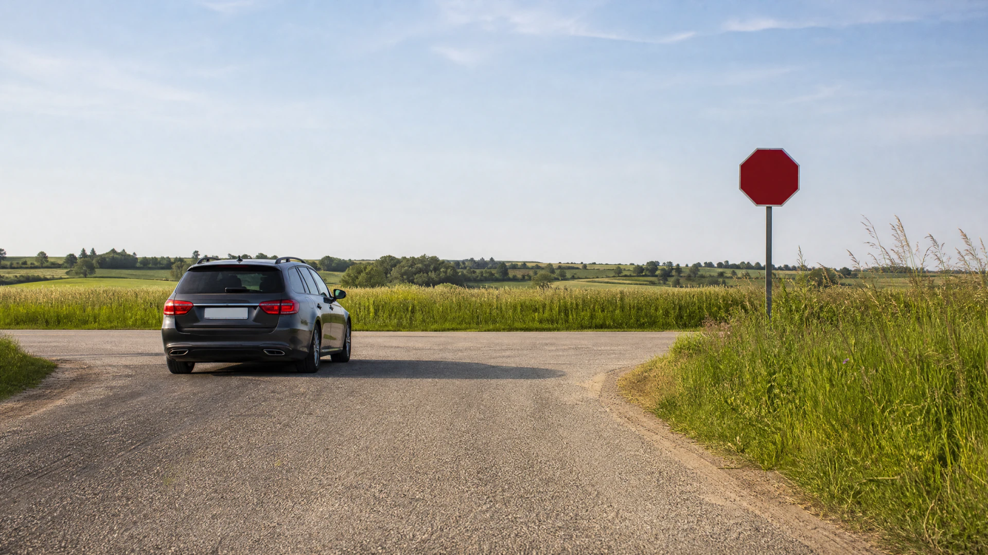 A dark gray station wagon sits at a rural crossroads beside a blank red octagonal sign mounted on a metal post.