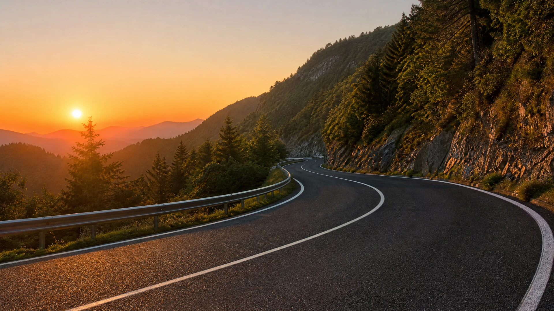 A paved mountain road with white lane markings curves along a rocky cliff face lined with conifer trees, set against a vivid orange sunset over distant mountain ridges.