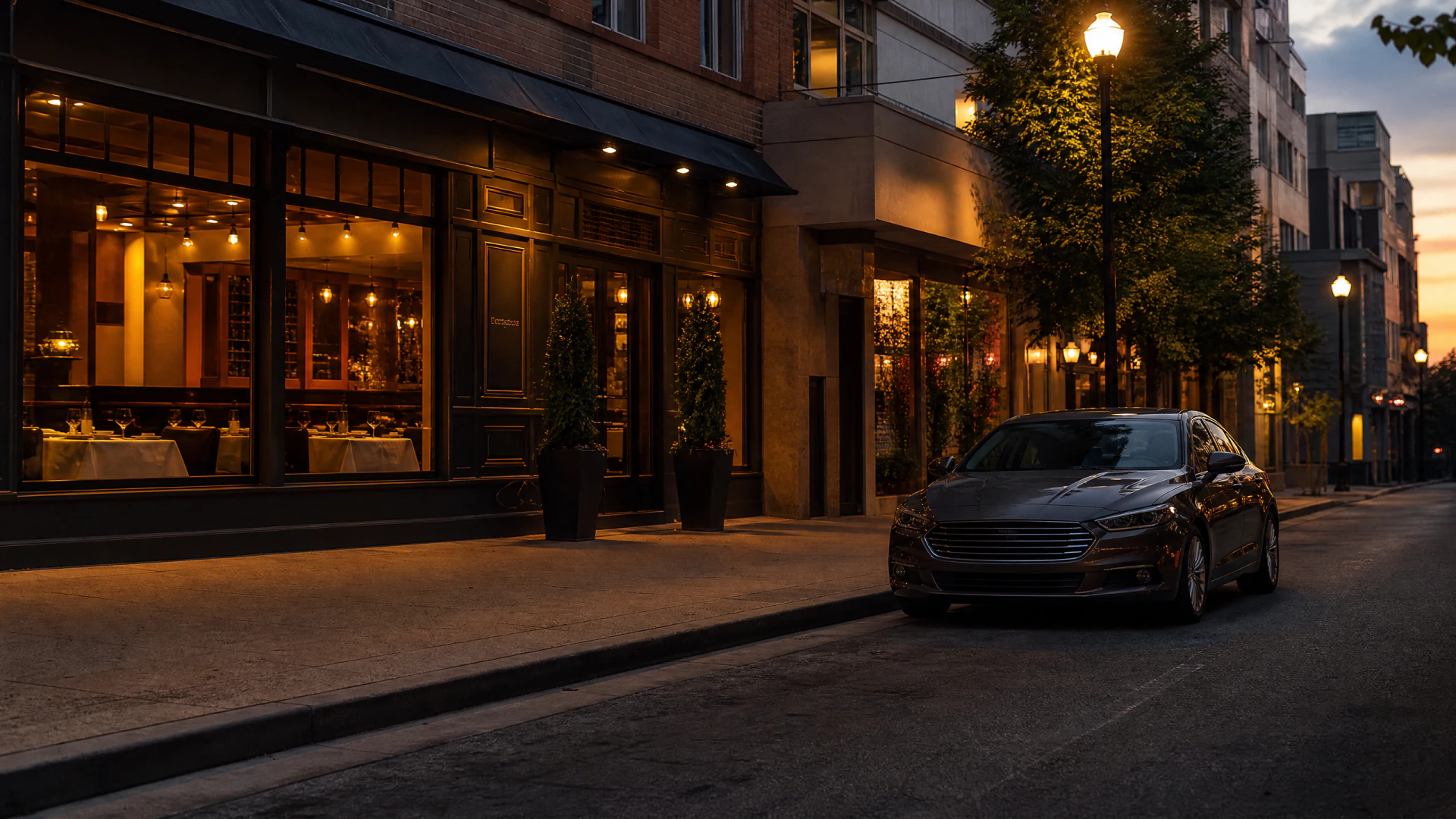 A brown sedan parked on an empty city street at dusk beside a warmly lit restaurant with large windows.